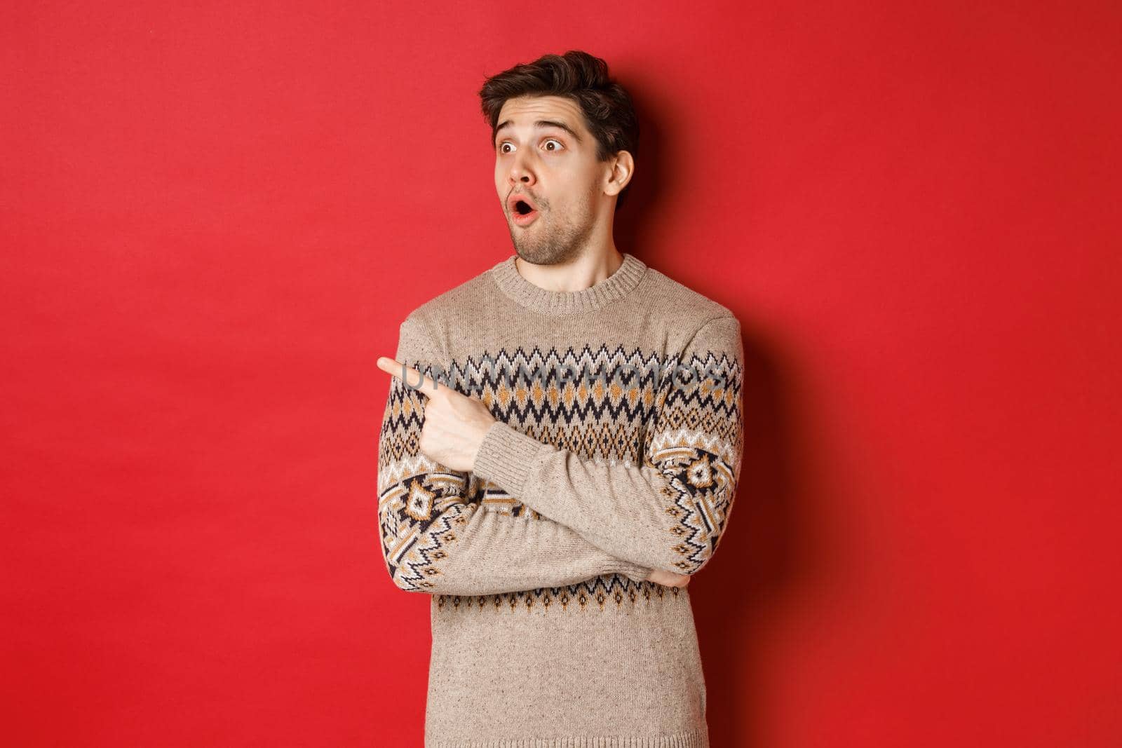 Portrait of attractive caucasian man celebrating new year, winter holidays, pointing finger and looking left surprised, showing christmas advertisement, standing over red background.
