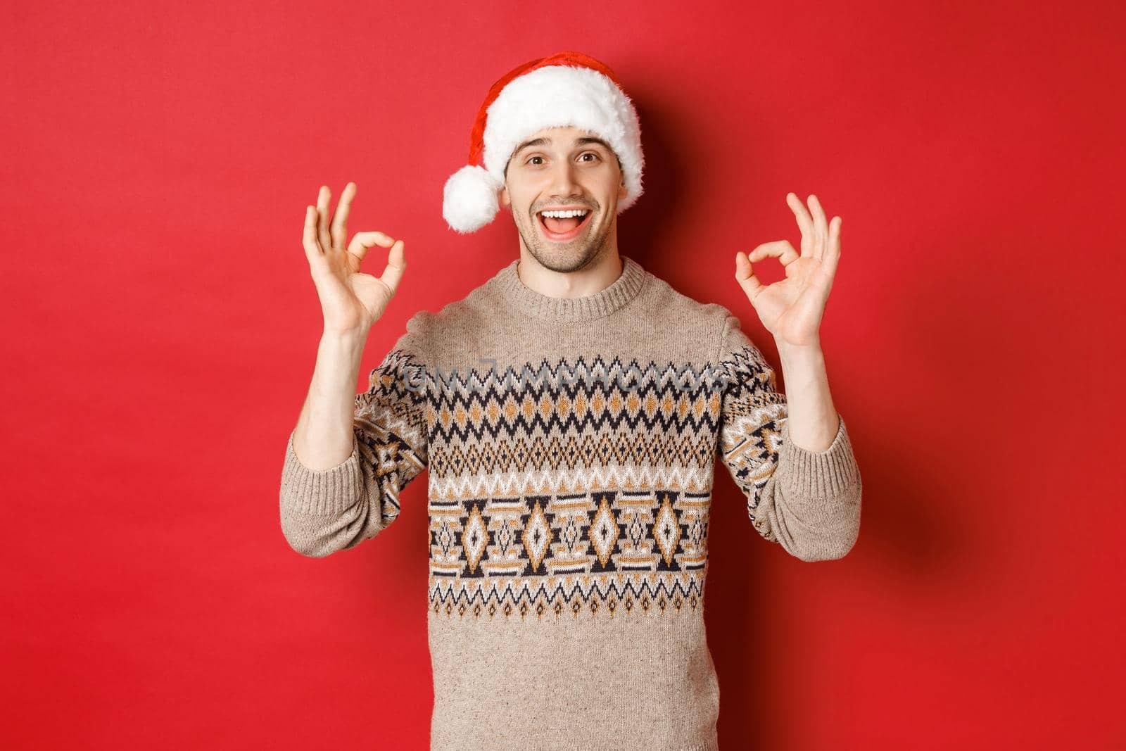 Portrait of happy and pleased handsome man, wearing winter sweater and santa hat, showing okay signs, praise good christmas party, standing over red background.