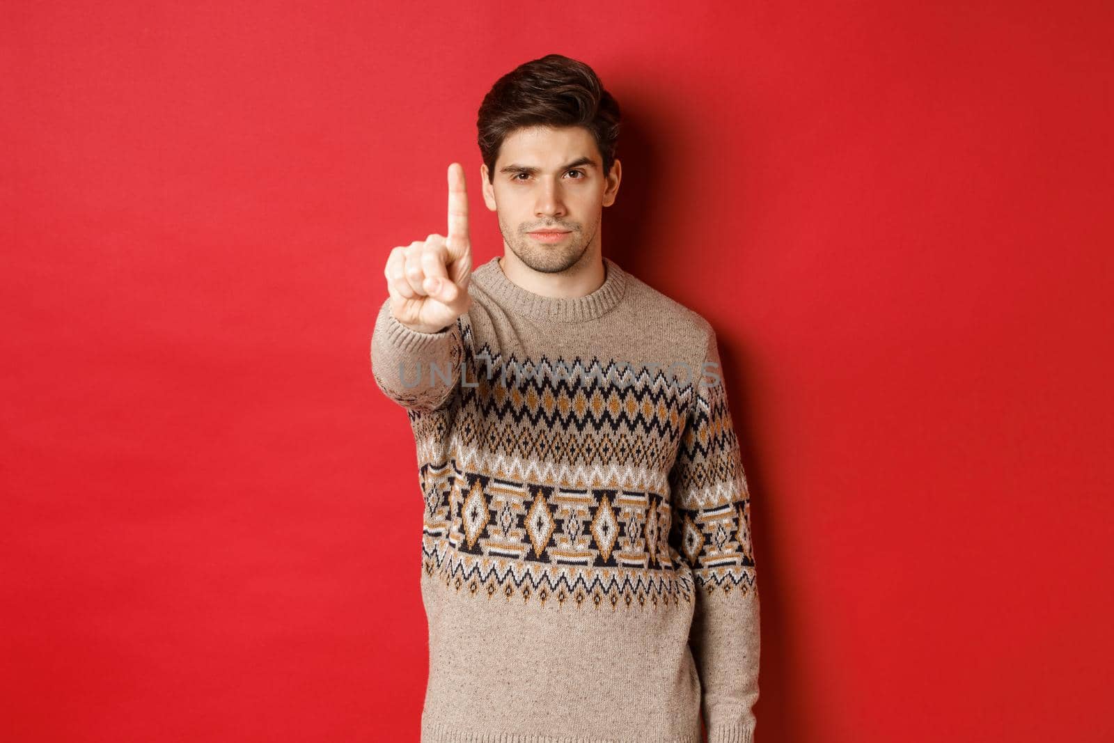 Image of serious handsome man extending one finger to stop you, telling no, give warning, standing over red background in winter sweater.