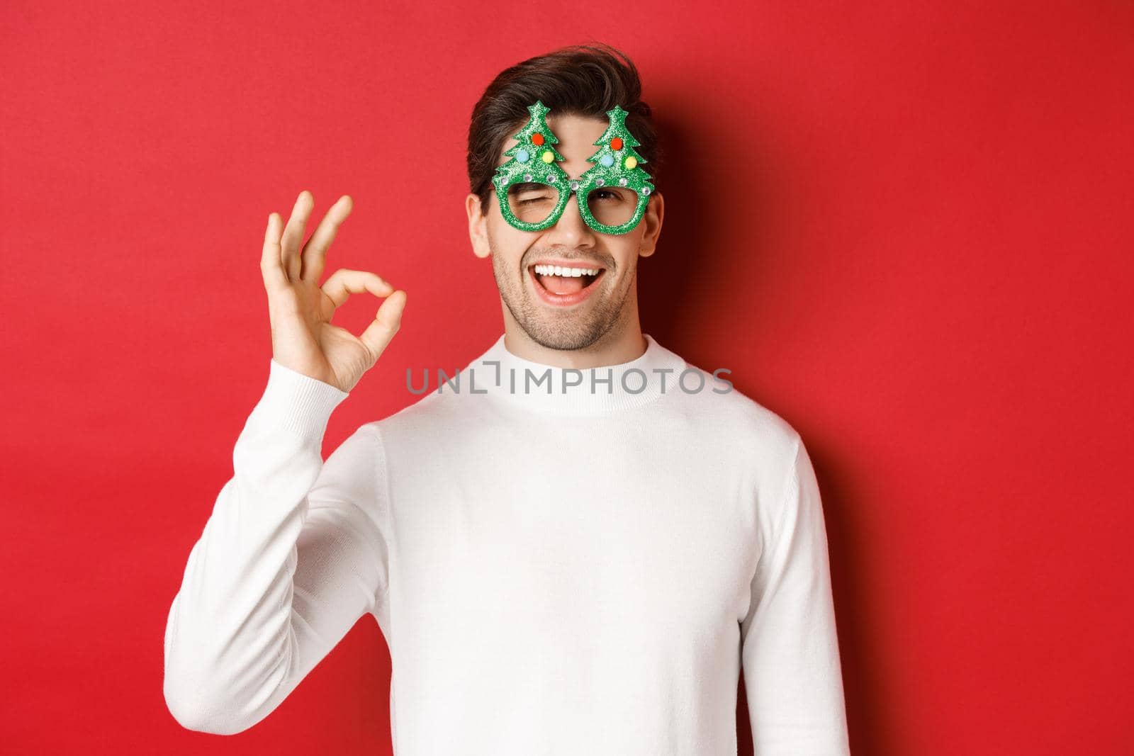 Portrait of handsome and cheeky guy, wearing party glasses and white sweater, showing okay sign and winking, wishing happy new year, standing over red background.