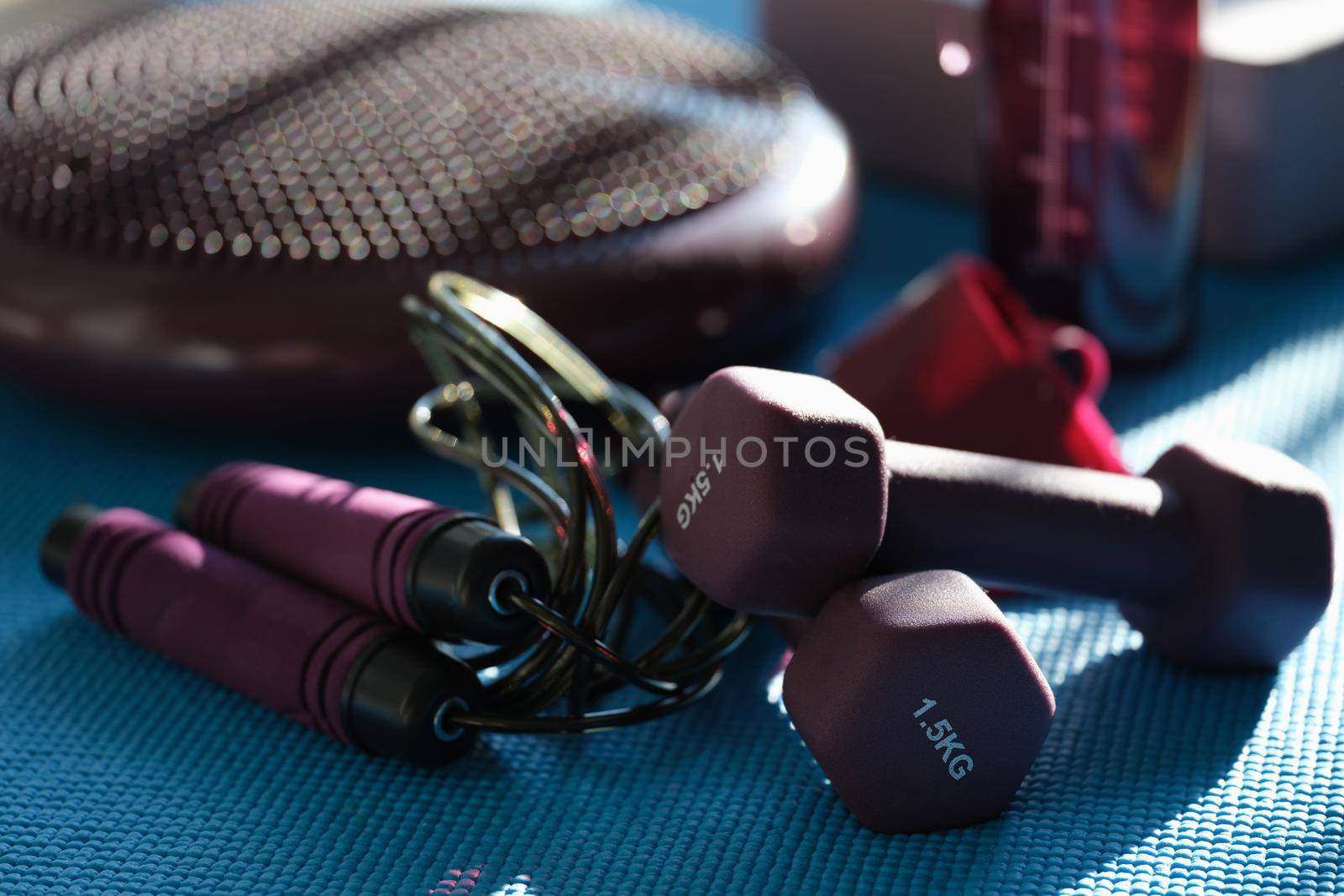Burgundy skipping rope dumbbells and massager on a blue sports mat, close-up, blurry. Fitness set, sun glare