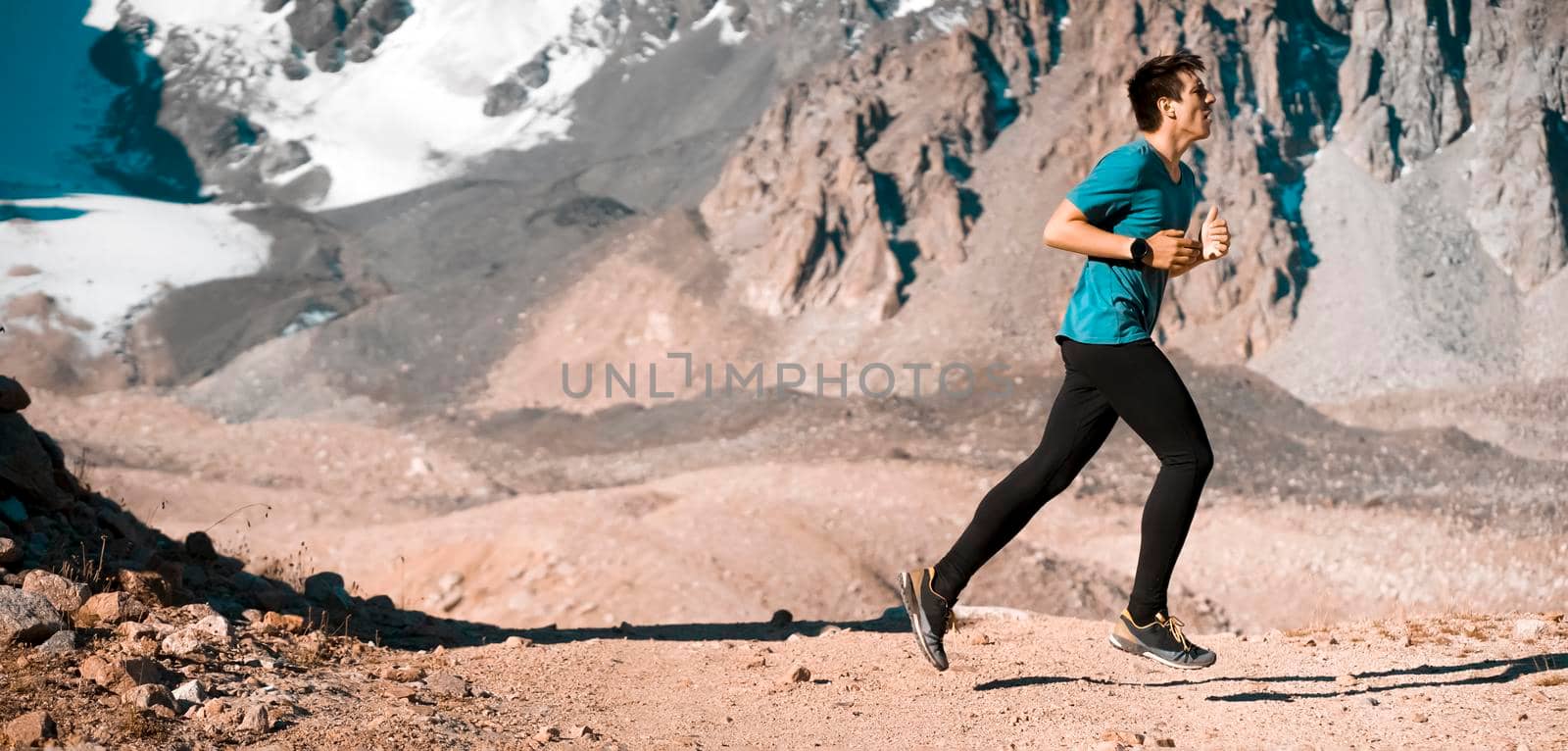 A young man runs along a trail in the snow-capped mountains, doing outdoor workout. The runner warms up, prepares for the race, leads a healthy active lifestyle.