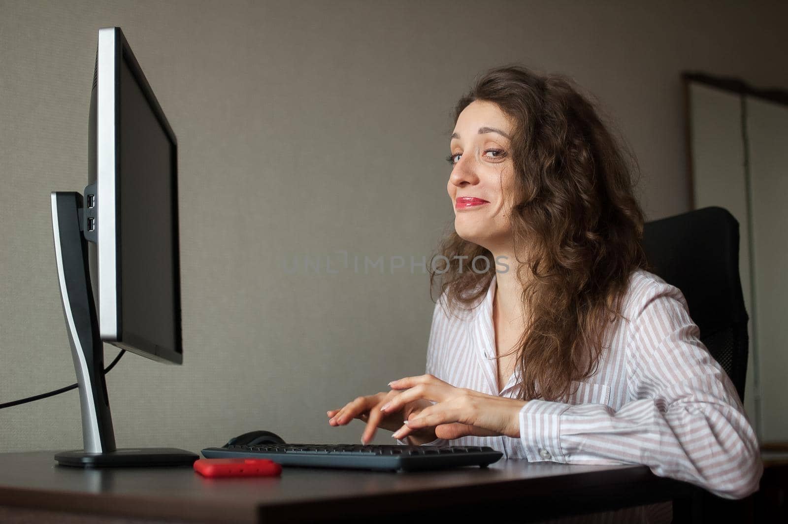 Young female office manager in white shirt and curly hair is sitting at the table and typing using keyboard, routine work, freelance by balinska_lv