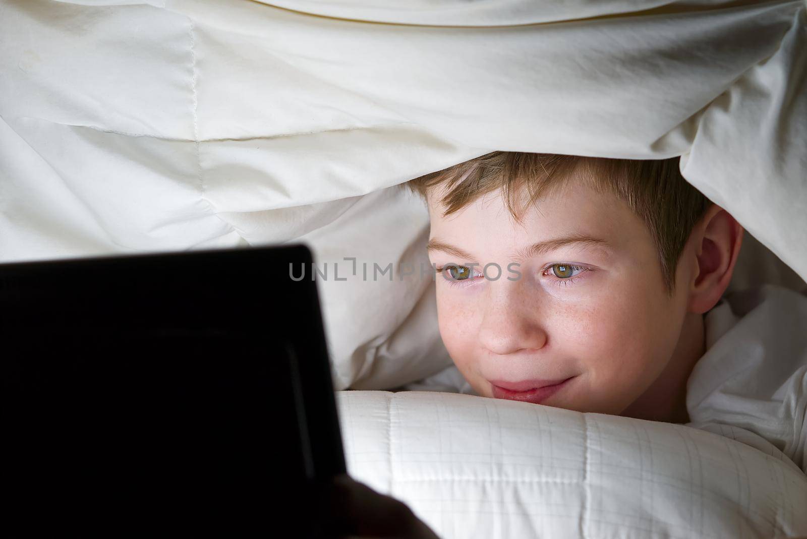 Quarantine. Distance learning. Online education. Happy boy in bed under blanket with tablet in dark. Child's face is illuminated by bright monitor. social media addiction. internet addiction.