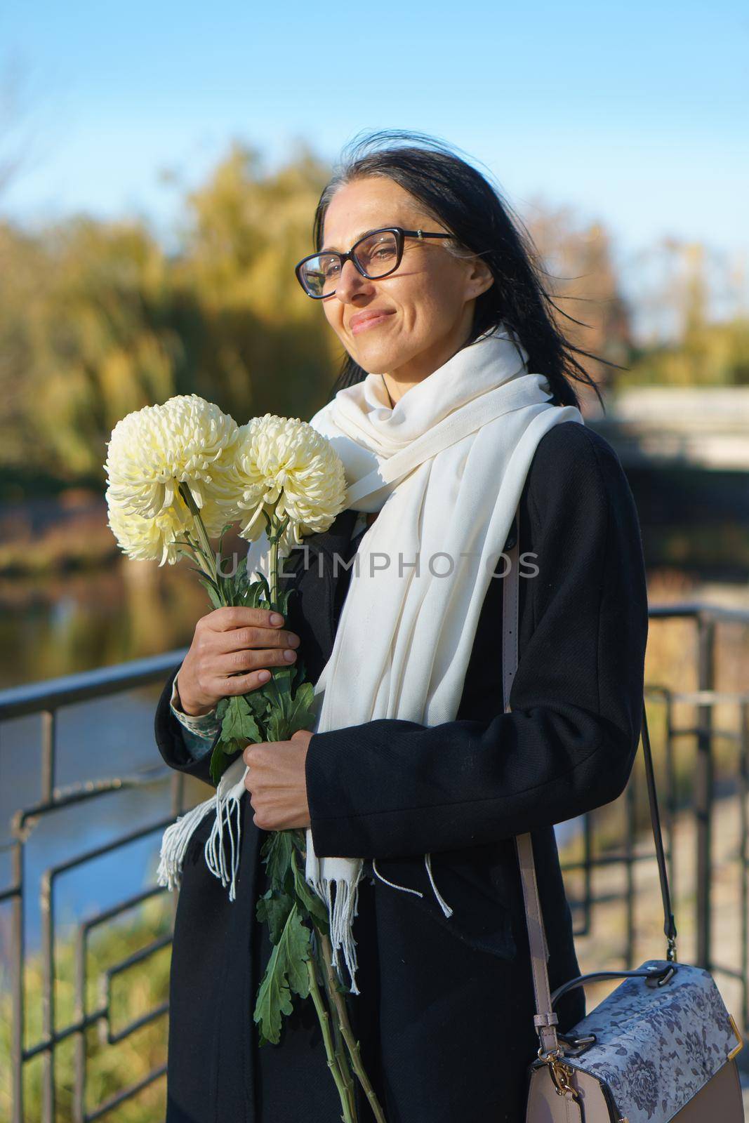 A beautiful middle-aged woman getting grey-haired in a dark coat in a spring town with a bouquet of flowers. by balinska_lv
