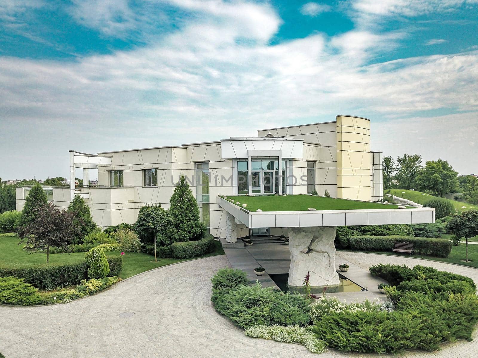 Luxurious modern country house with roofed porch at main entrance and massive decorative stone column supporting roof, surrounded by green parkland in cloudy summer day