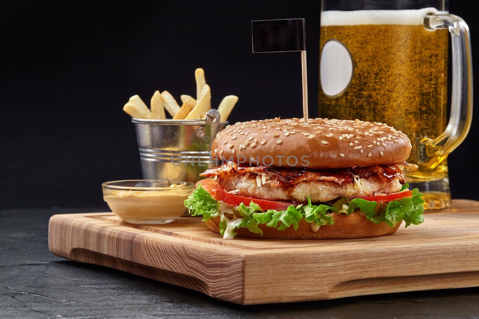 Tasty burger with chicken patty, fried bacon, grated parmesan and vegetables in sesame bun served with glass of foamy beer, crispy fries and aioli sauce on wooden board. Popular fast food dish