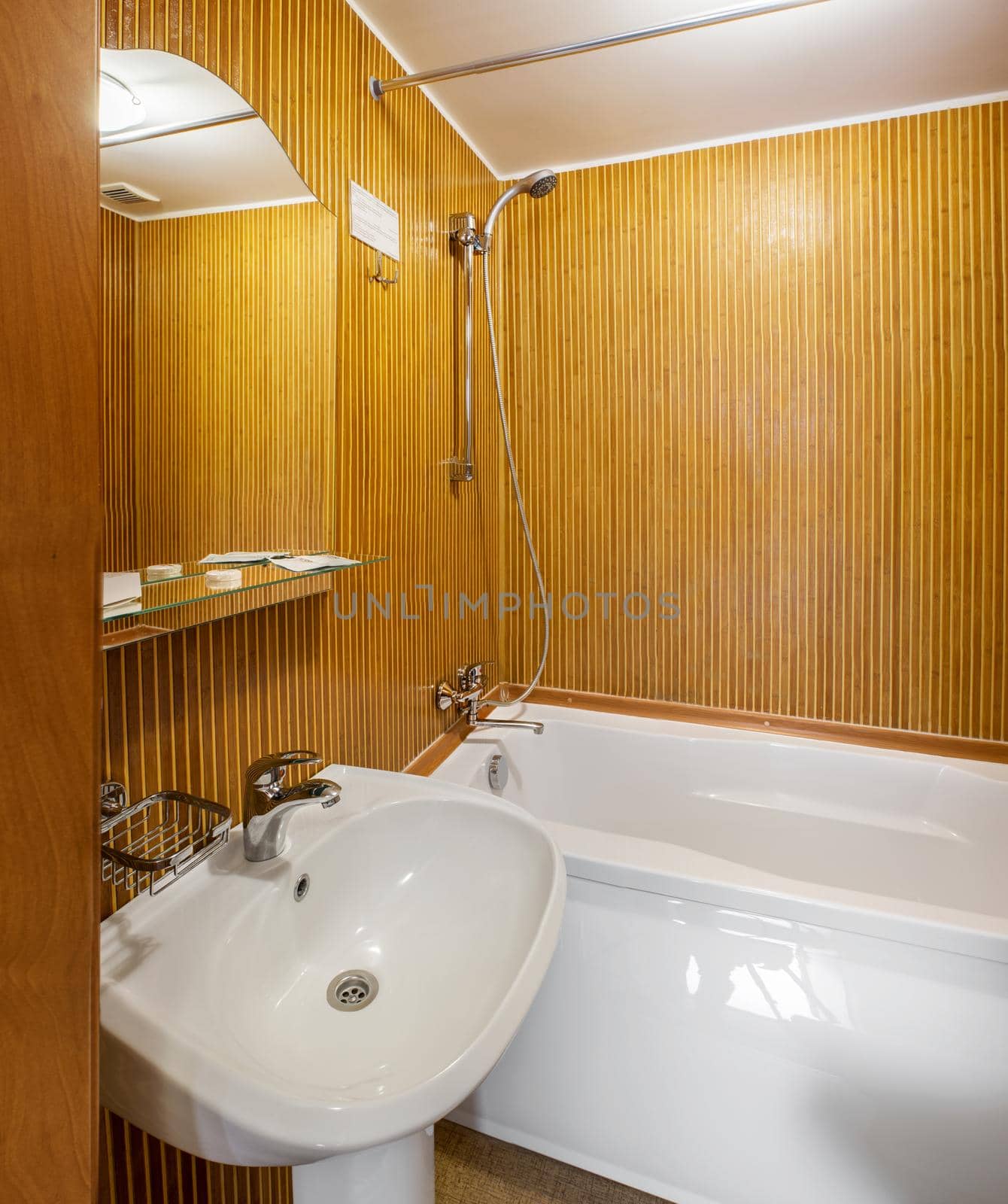 Interior of small bathroom in hotel room with walls faced with wooden slats, equipped with white wash basin and acrylic bathtub with chrome fixture and mirror