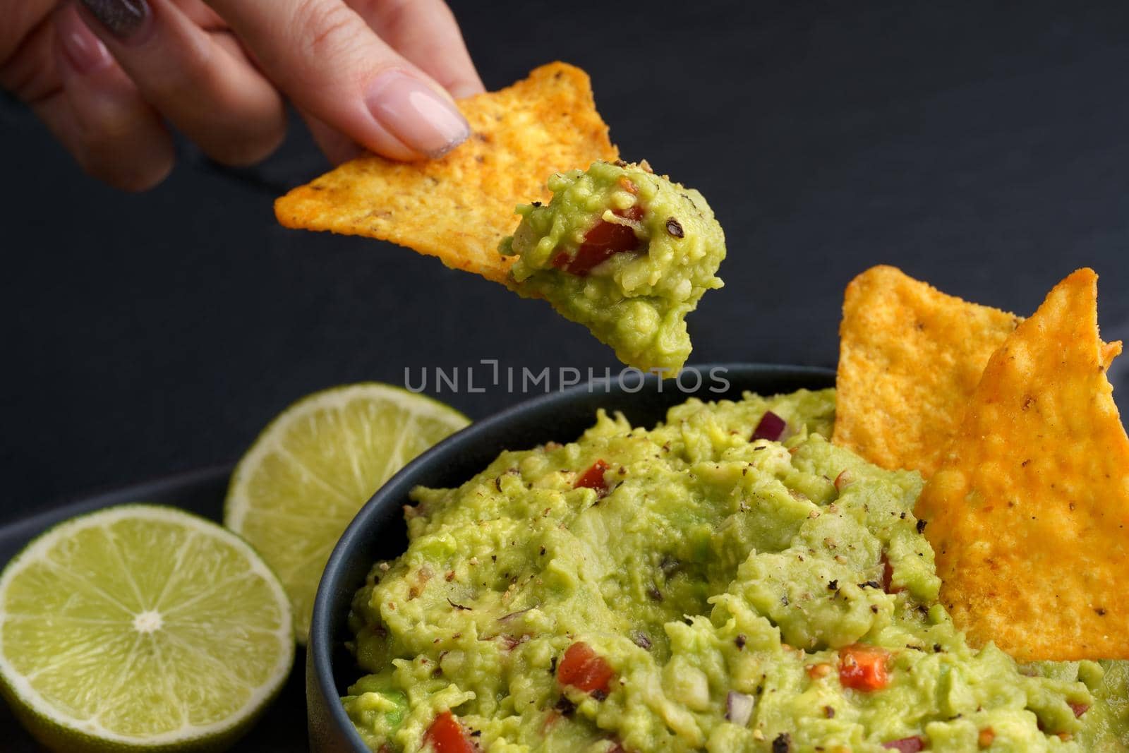 Woman hand holding tortilla chip or nachos with tasty guacamole dip on a black background by DariaKulkova