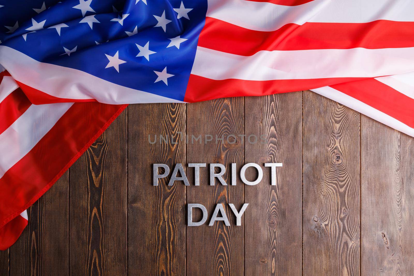 the words patriot day laid with silver metal letters on wooden board surface with crumpled usa flag above, in flat lay high angle directly above view