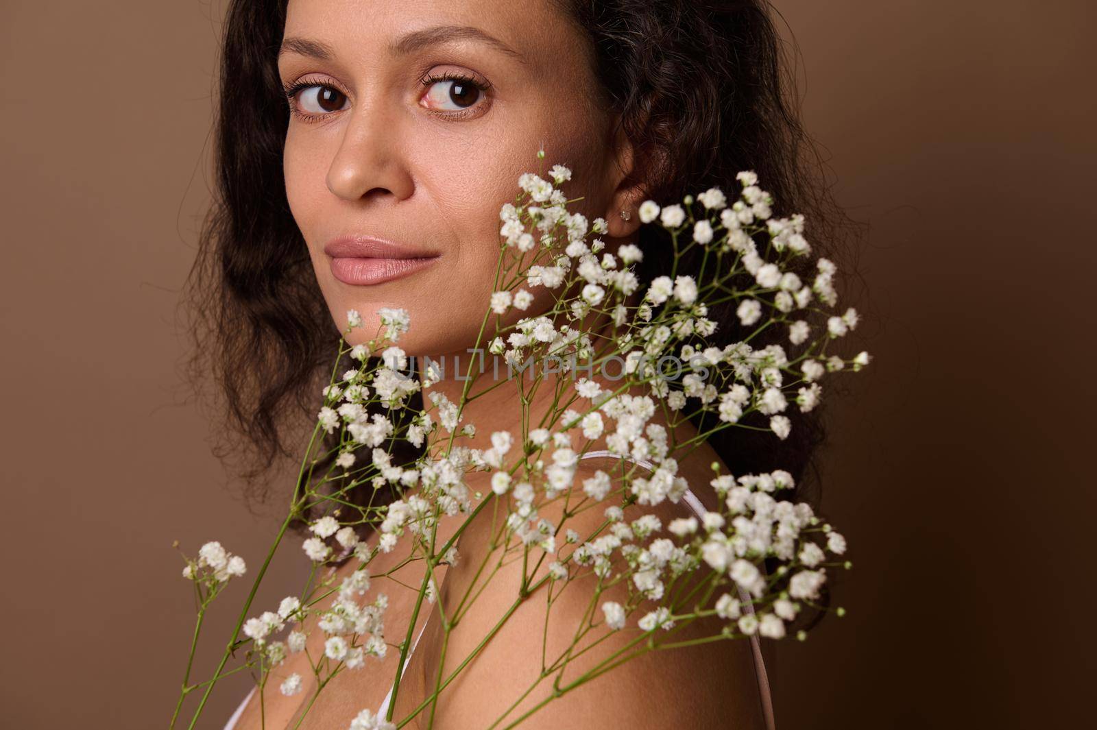 Close-up portrait. Beautiful woman with white gypsophila sprig standing three-quarters against brown background looking confidently at camera. Body care, positivity, femininity, Women's day concept