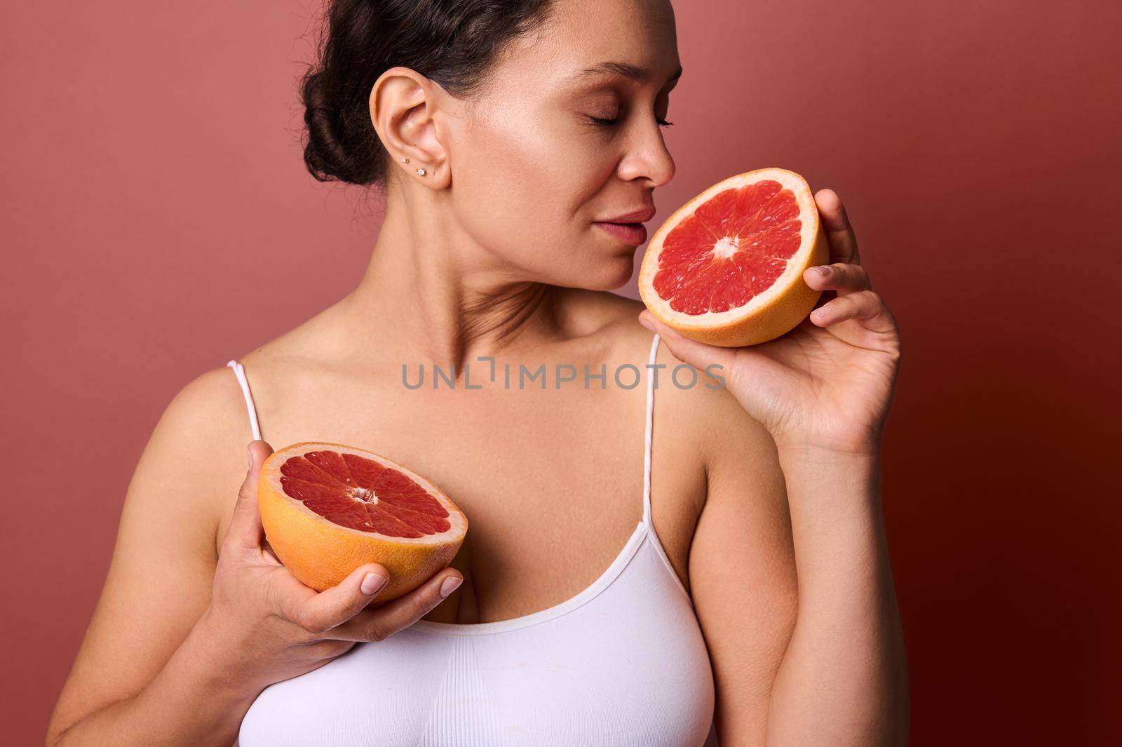 Middle aged woman holding grapefruit halves in her hands, enjoying their aroma, isolated on coral background with advertising space. Natural organic raw ingredients and healthcare and medicine concept by artgf
