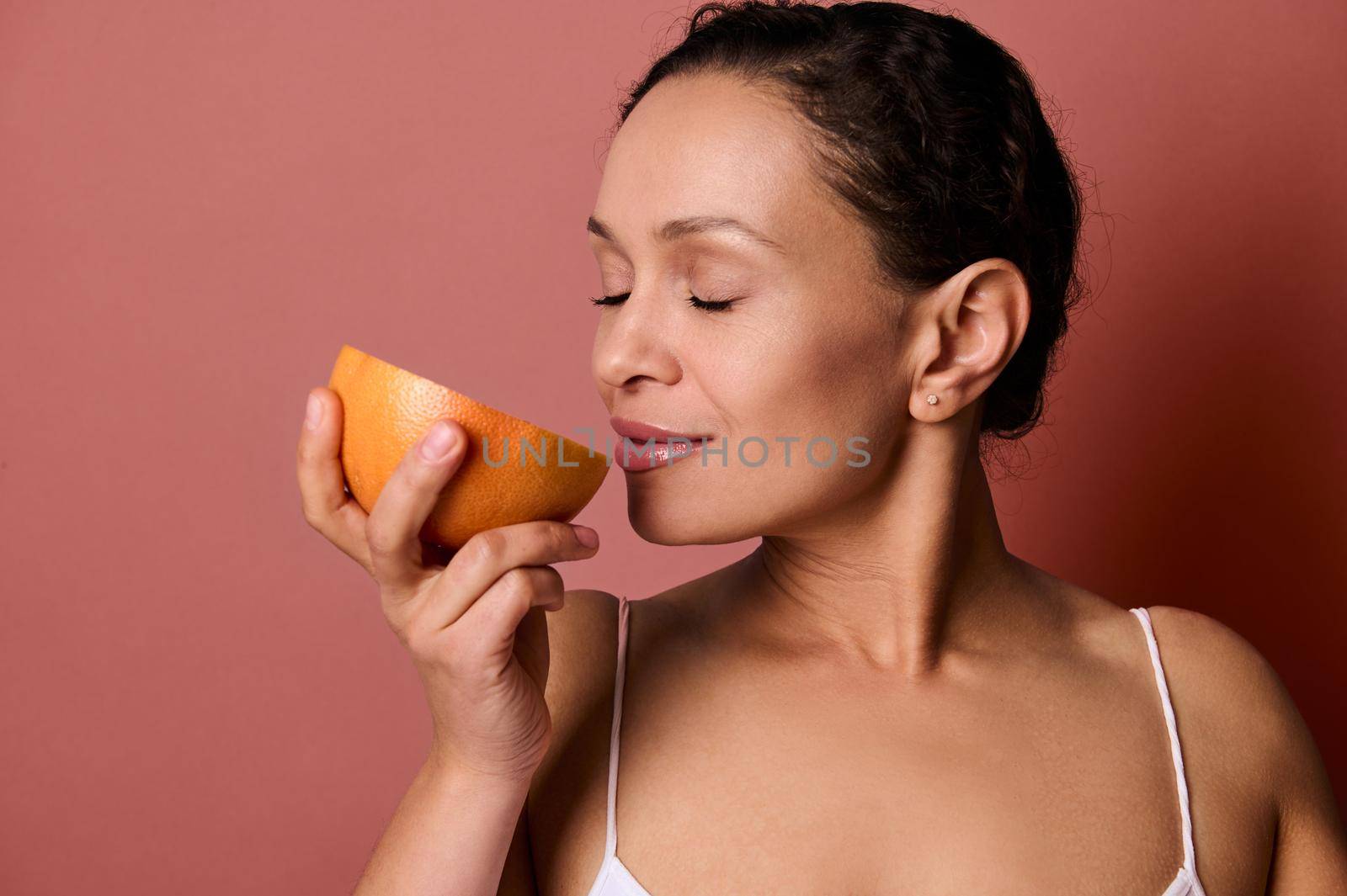 Close-up of dark-haired pretty woman smelling the scent of fresh juicy red grapefruit, posing with closed eyes against coral background with copy space. Health, body positivity and skin care concept.