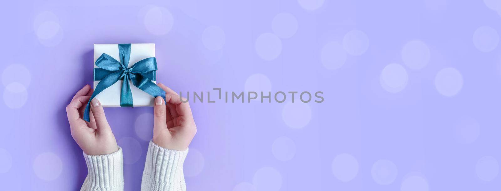 Banner of Female Hands in sweater holding gift in white wrapping paper on violet background. St. Valentines Day, love, tenderness, friendship, Birthday, Christmas. Festive wallpaper. Color 2022