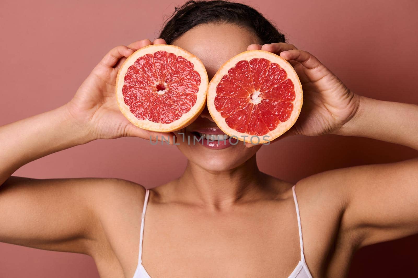 Charming young woman covering her eyes with two halves of fresh juicy grapefruit, smiling with beautiful toothy smile, posing against pastel coral background. Skin and body care concept. Copy space by artgf