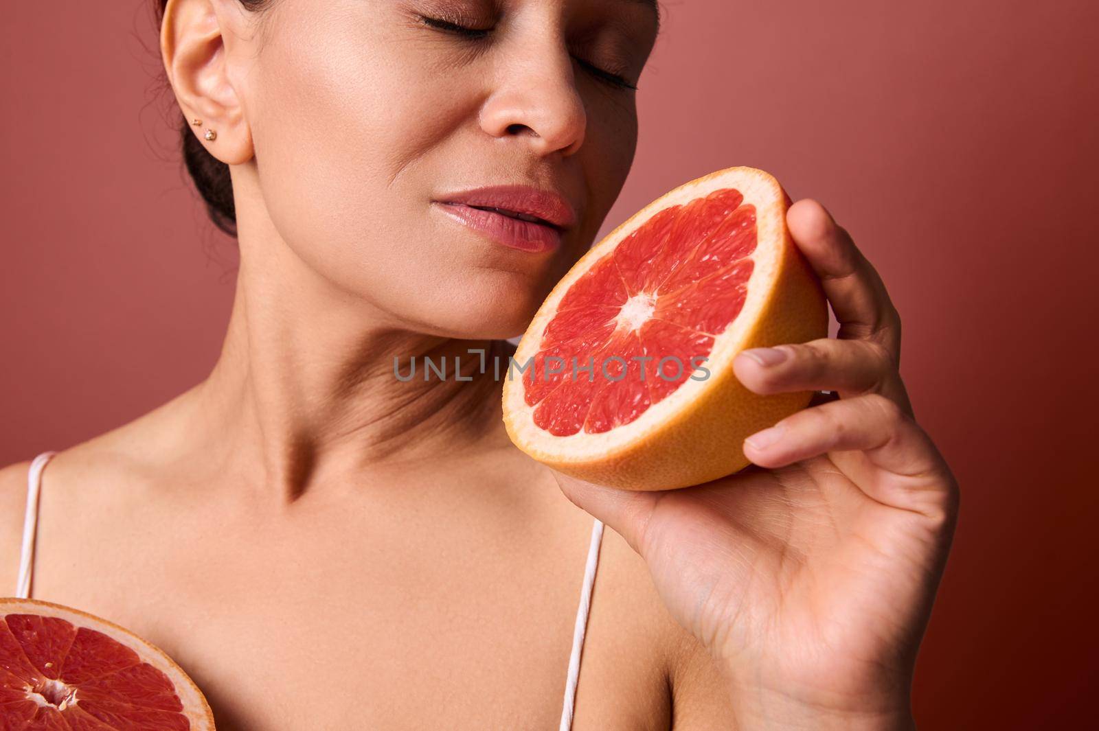 Close-up of middle aged beautiful woman with healthy glow perfect smooth skin holding grapefruit halves near her face. Natural cosmetics, skin care, wellness, beauty treatment, cosmetology concept
