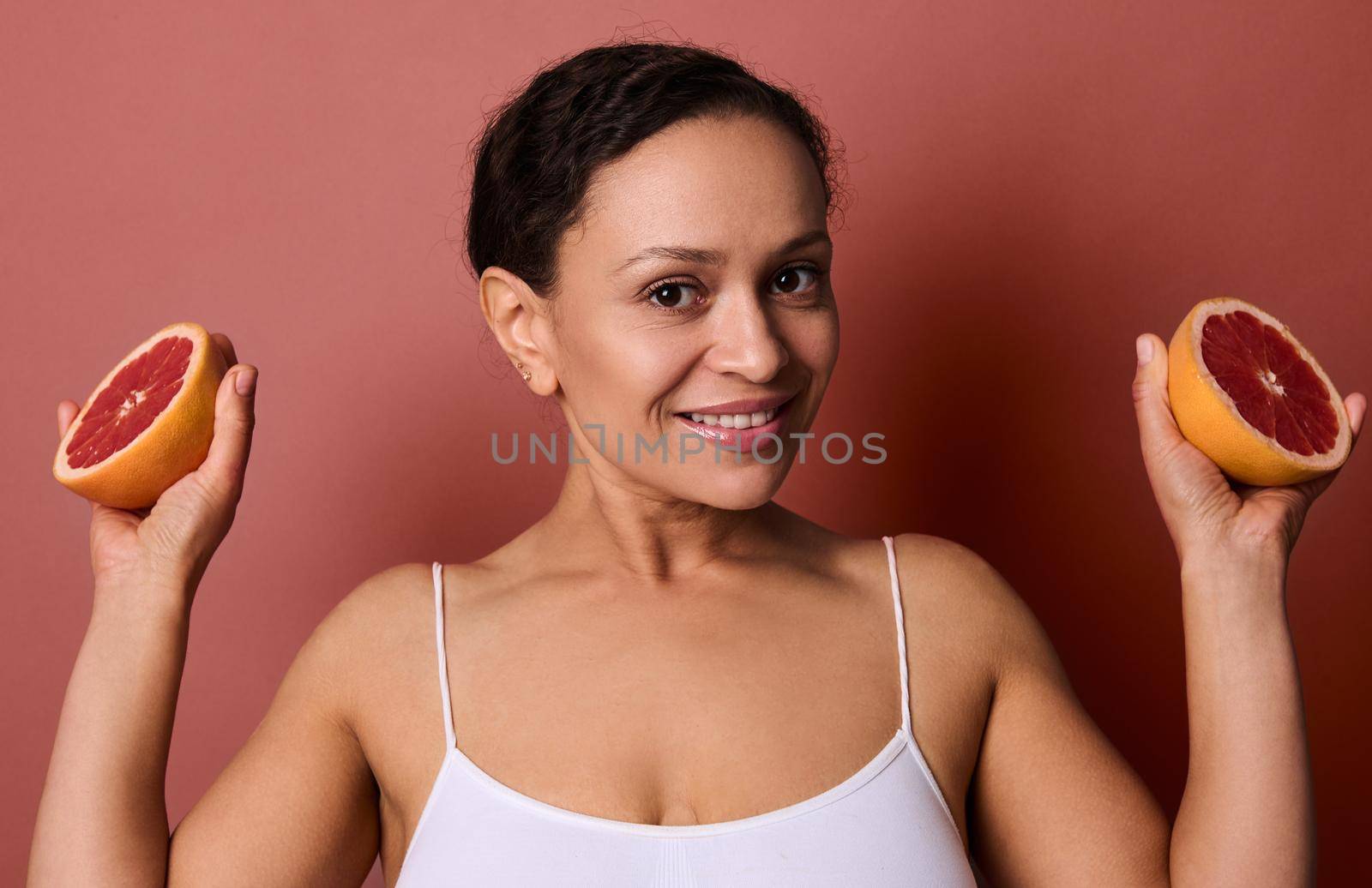 Charming middle aged African woman in white top smiles looking at camera, holding halves of red juicy fresh grapefruit in her hands, isolated over coral background with copy space for ads. by artgf