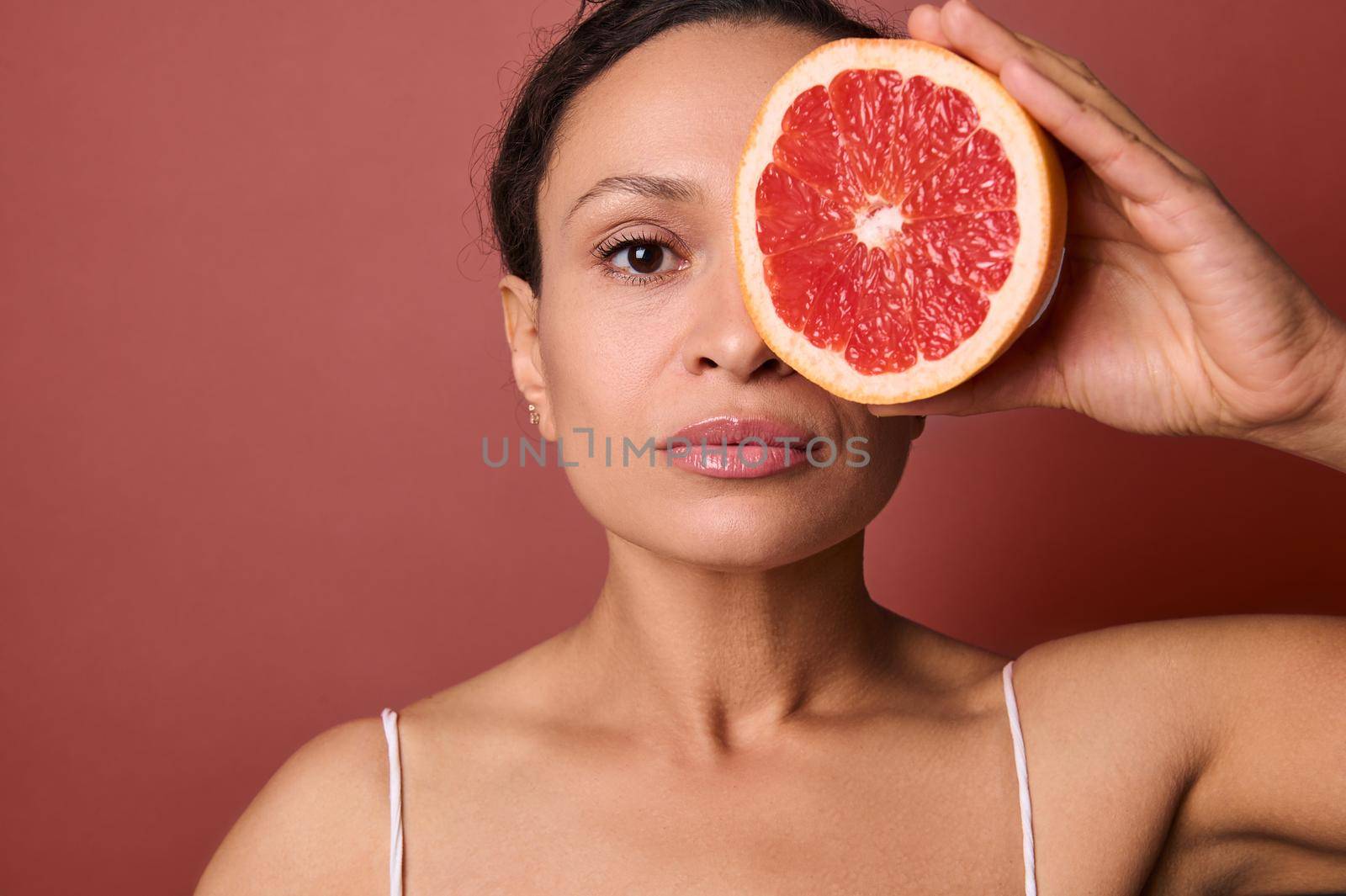 Attractive woman with perfect healthy skin holds a grapefruit in front of her eye, covering with it half of her beautiful face. Using natural organic plant based ingredients for skin care cosmetics by artgf