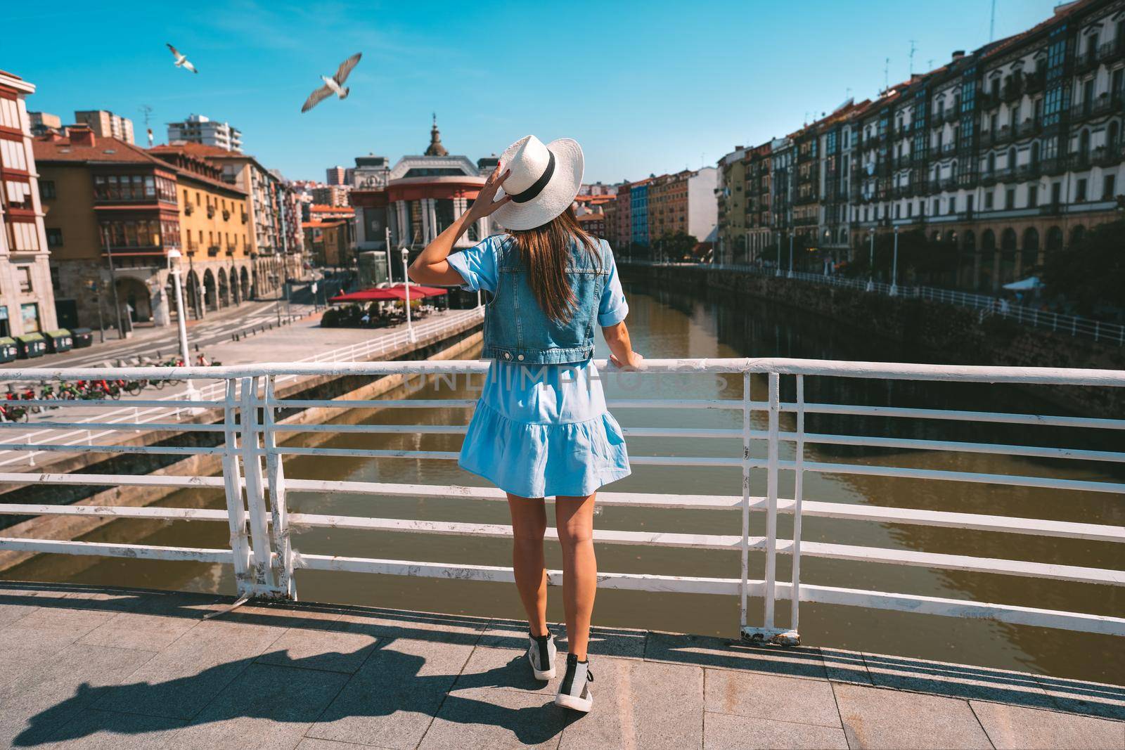 Rear view of young tourist woman in white sun hat walking in Bilbao city. Summer holiday vacation in Spain. High quality photo