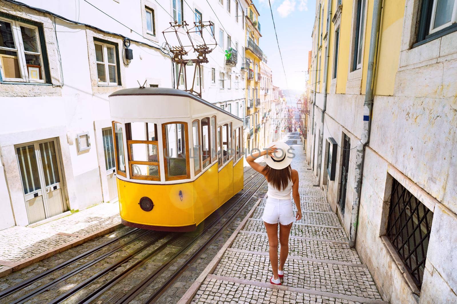 Woman tourist walking in narrow streets of Lisbon city old town. Famous retro yellow funicular tram on a sunny summer day. Tourist attraction. Vacation and travel concept. High quality photo