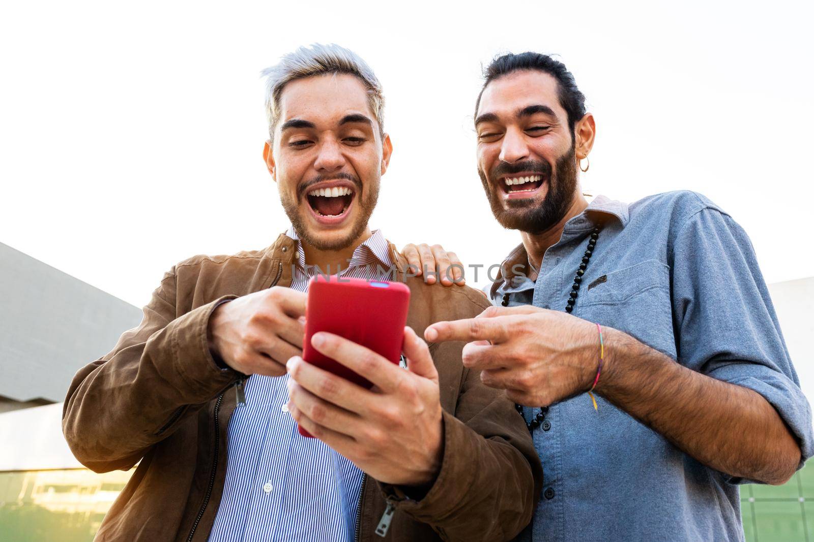 Young male gay couple look at mobile phone. Caucasian men watch funny videos on cellphone together. LGBT and technology concept.