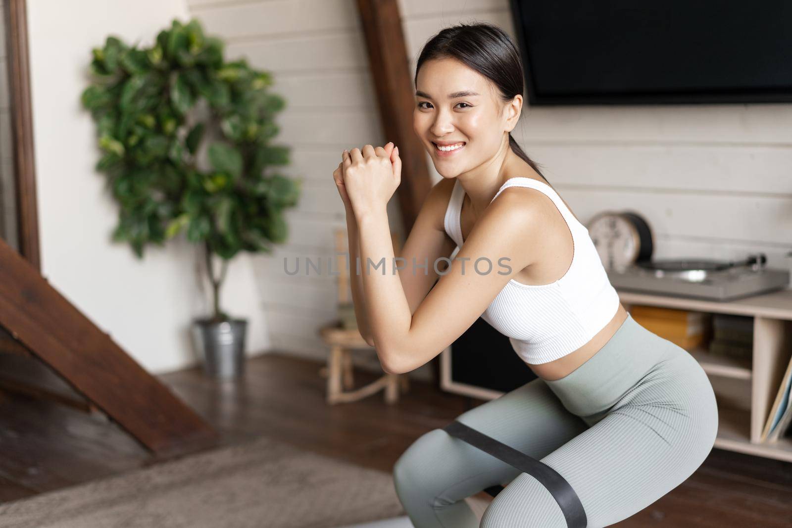 Cute asian fitness girl at home doing workout, squats with stretching elastic rope on her legs, exercising for fit and healthy body.