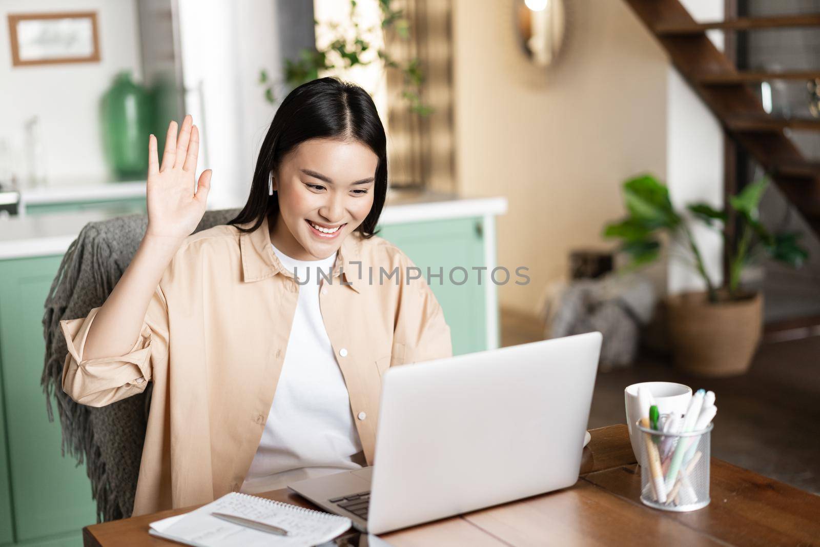 Smiling asian woman waving hello at video chat on laptop, sitting at home, studying remotely in e-learning school.