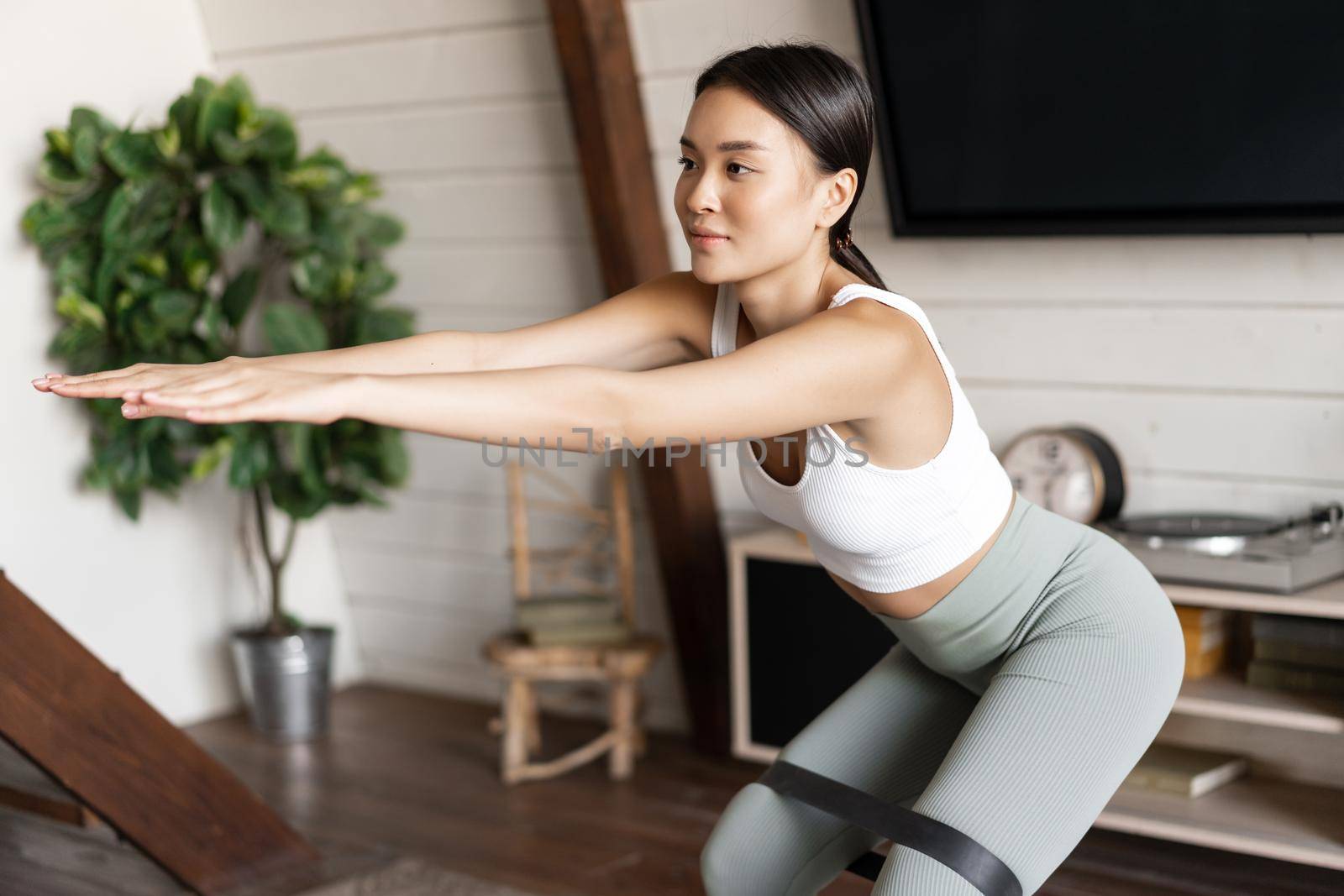 Cute asian fitness girl at home doing workout, squats with stretching elastic rope on her legs, exercising for fit and healthy body.