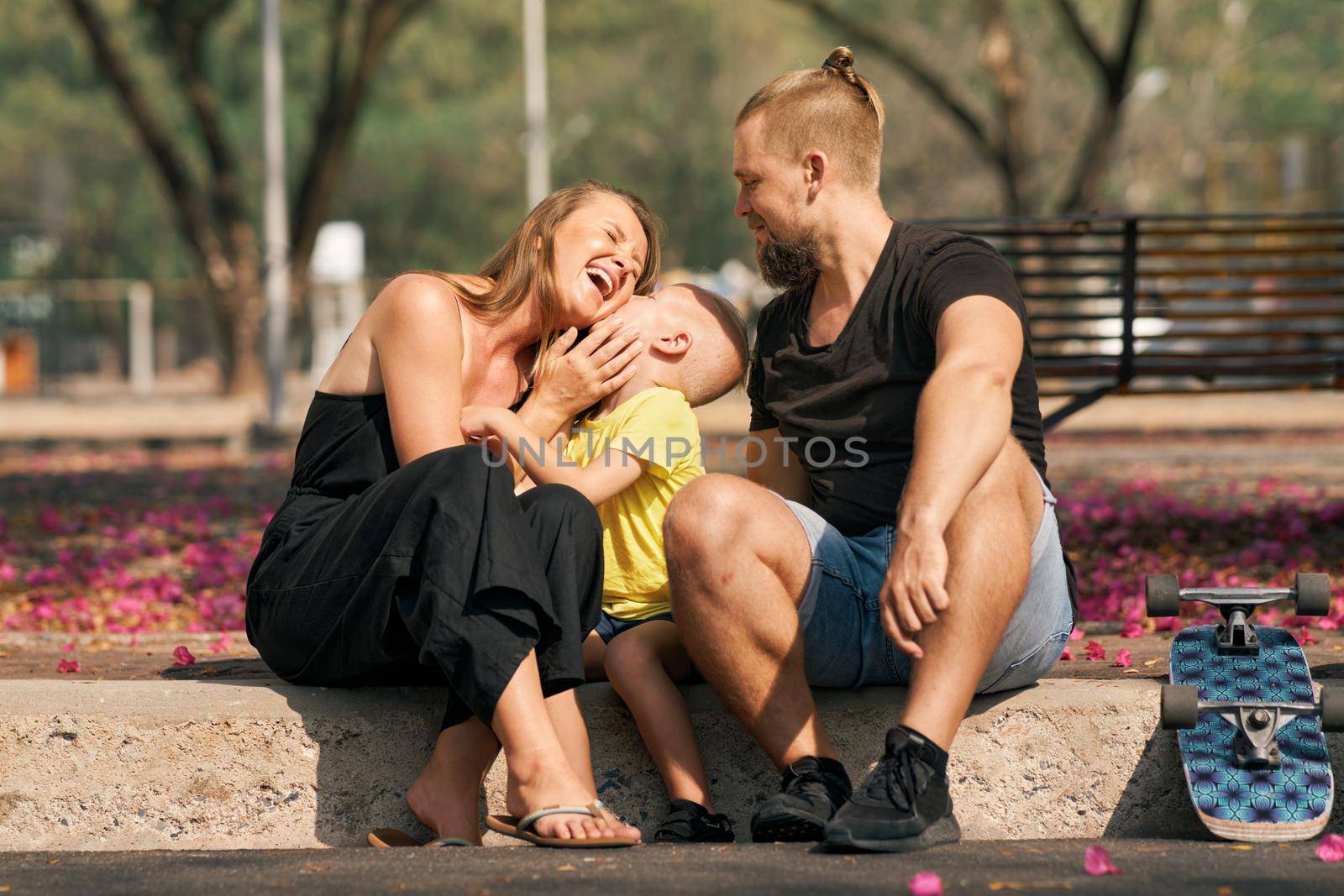 Closeup portrait father, mother is hugging their little son while sitting in skate park with blossom trees around. Concept of happy family love and care. Outdoor leisure and activity.