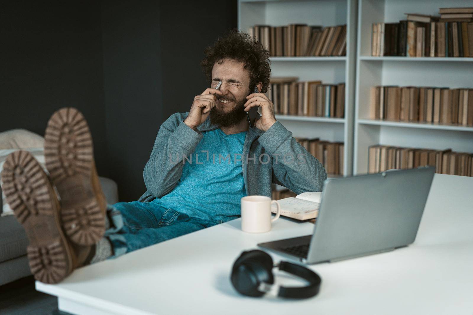 Male Model Poses with Feet on the Table and Smartphone in his Hand.There are Laptop, Mug of Tea, Diary and Earphones on the White Desk . Shelves Background. Close-up . High quality photo
