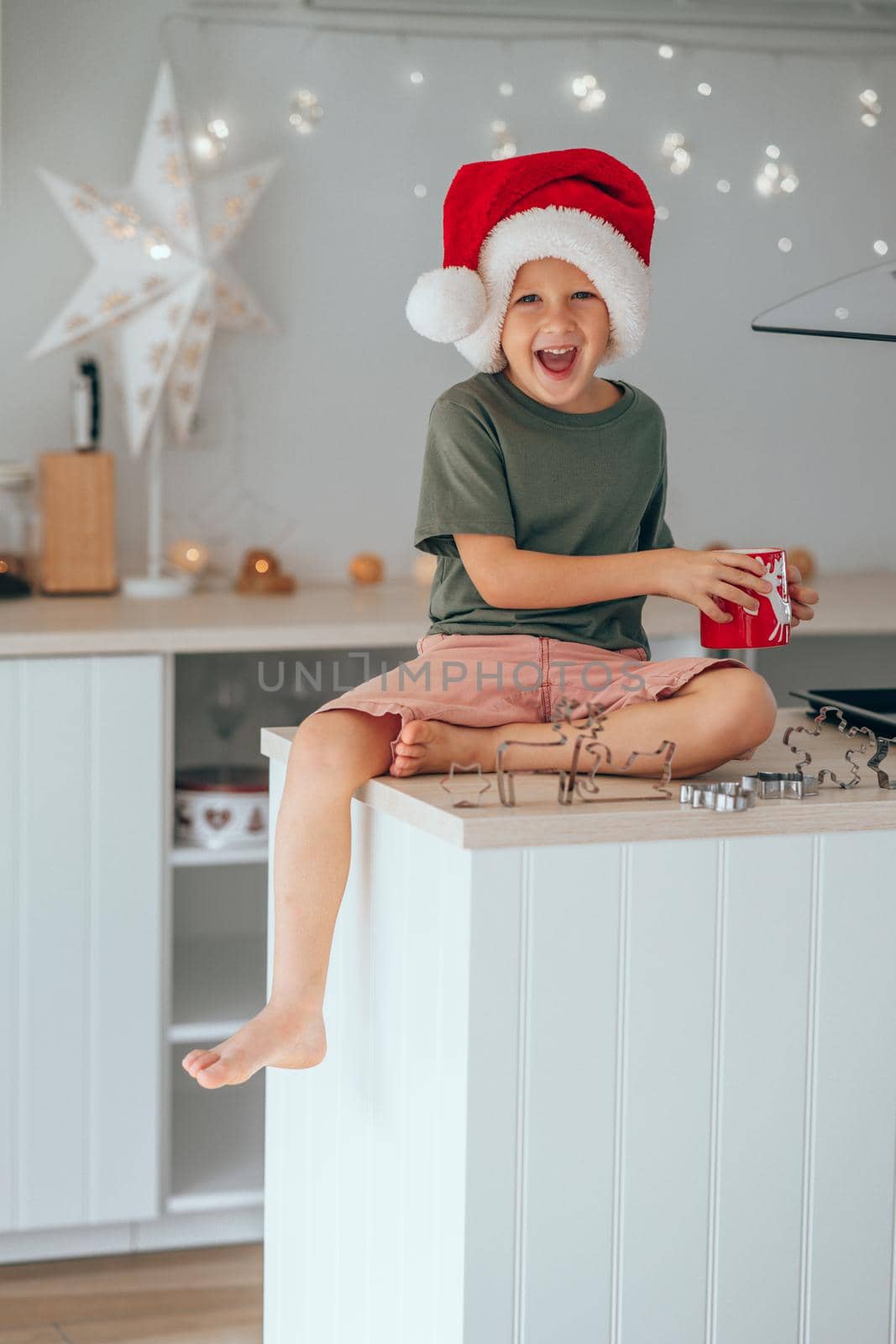 Happy little boy in a Santa hat and drinking cocoa milk sitting on the kitchen table while baking Christmas cookies for Christmas Eve. Decorated kitchen with garland to celebrate of Christmas holiday.