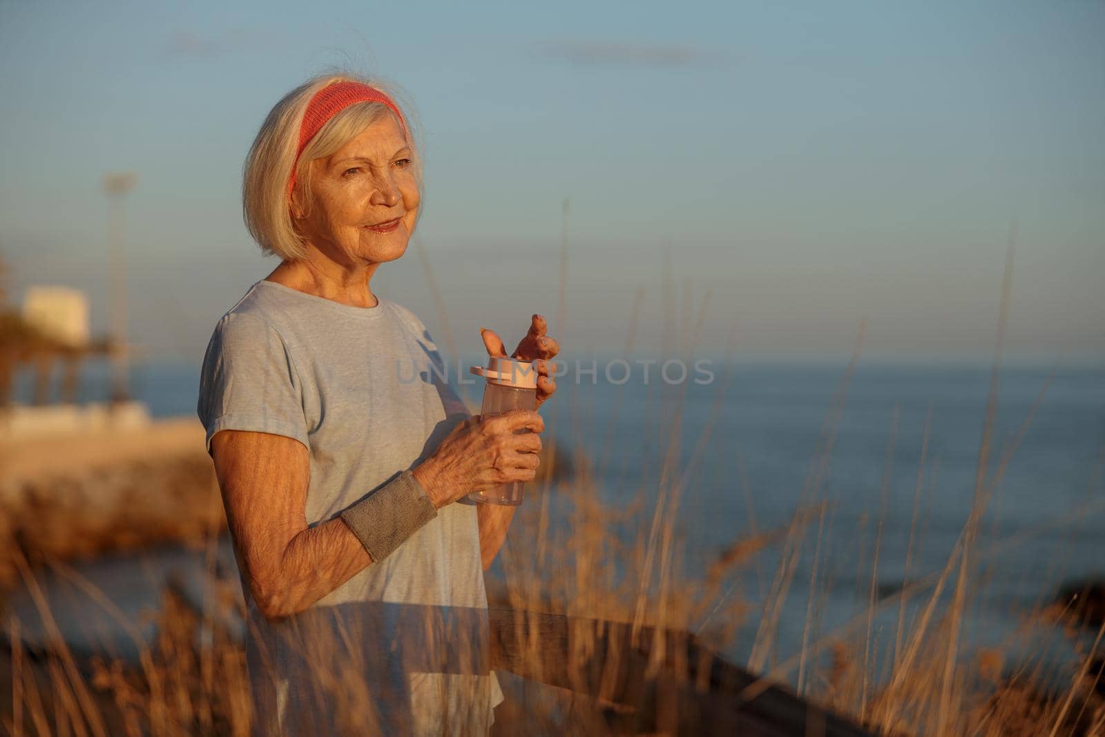 Relaxed elderly lady wearing sports clothes holding bottle with water and breathing fresh air, standing in autumn nature at sunset