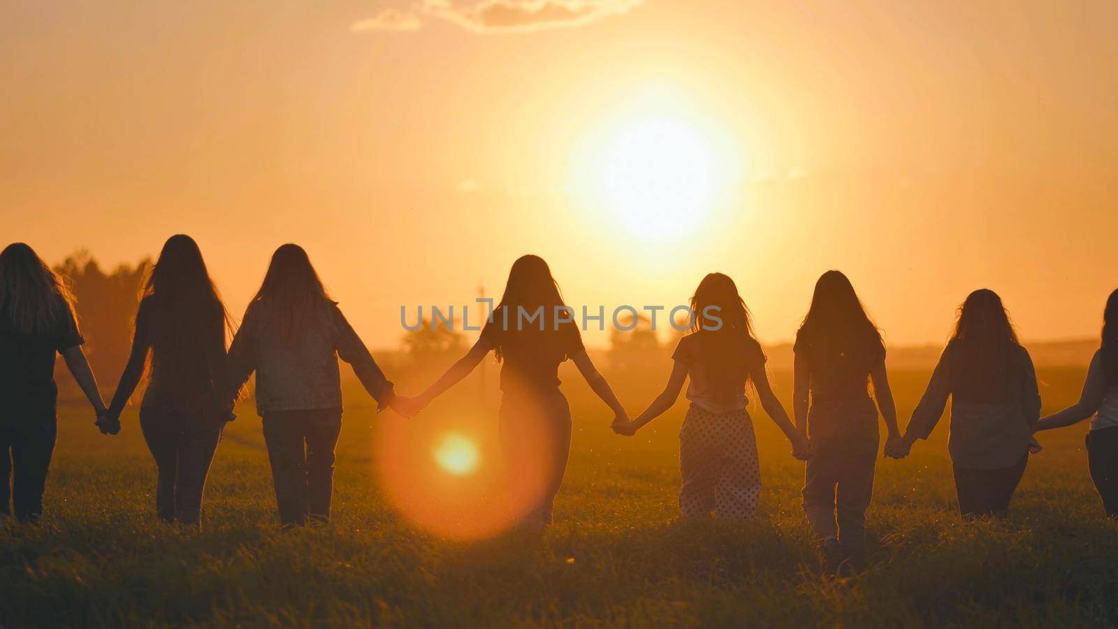 A group of girls walk towards the sun at sunset holding hands. by DovidPro