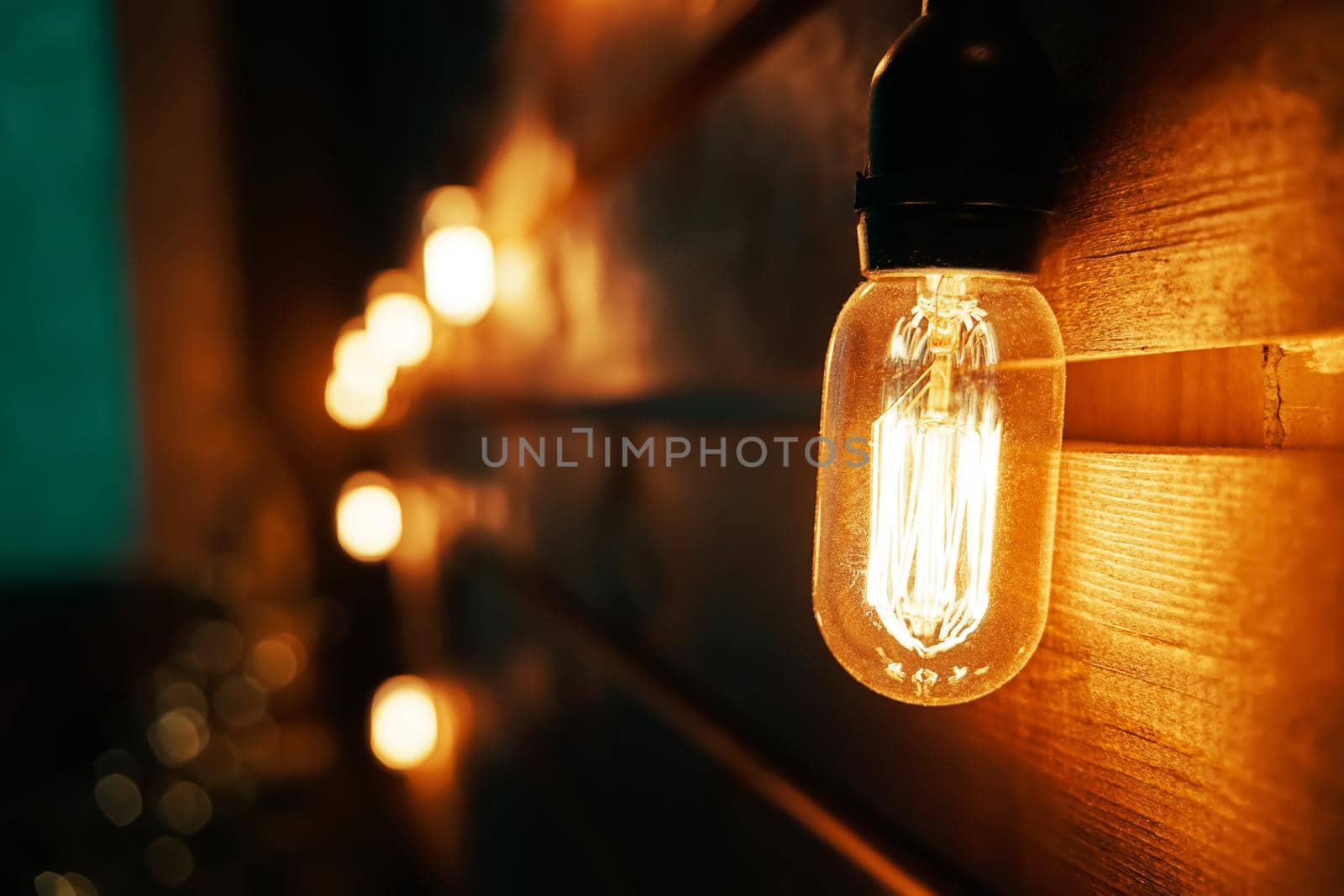 Holiday interior decorating. Antique incandescent Edison type bulb on wooden texture wall. Blurred warm garland light on background. Christmas night party mood. Copy space for congratulations.