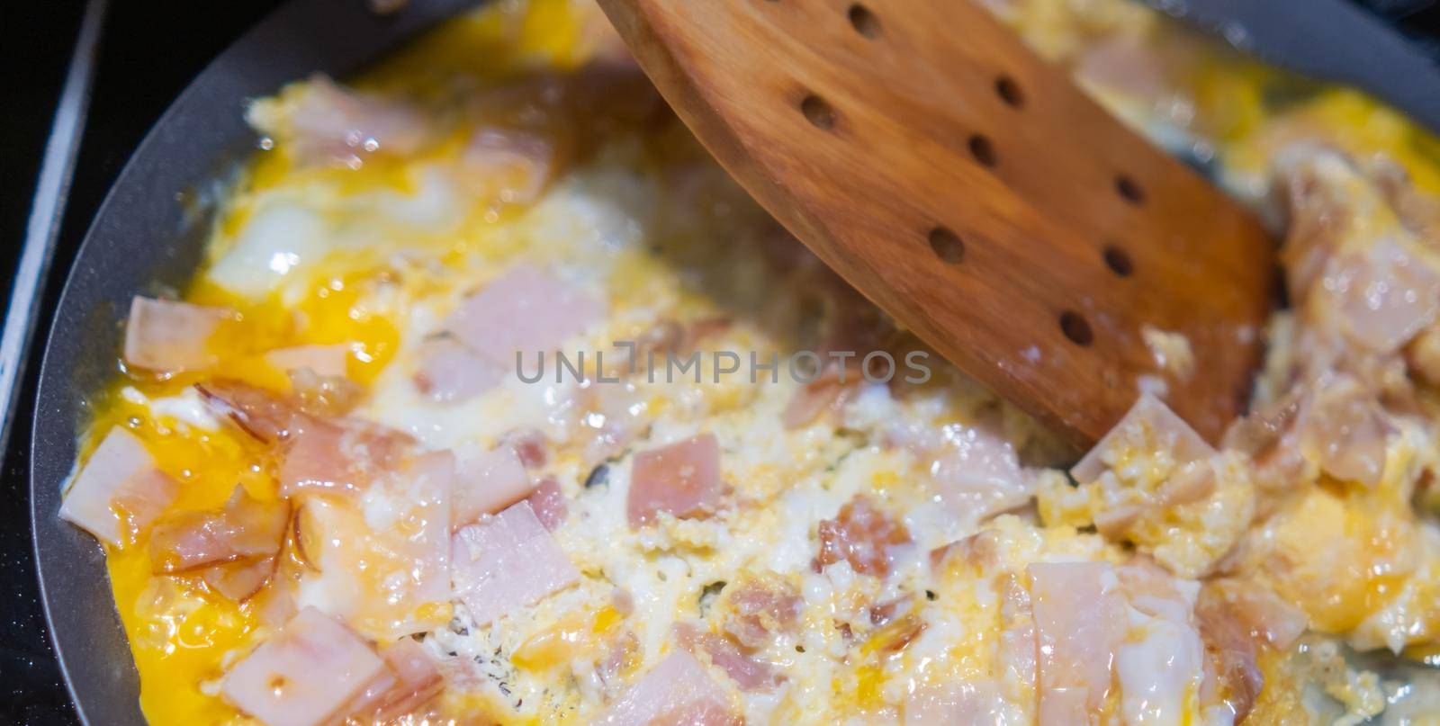 Close-up of wooden spatula mixing chopped turkey ham and raw scrambled eggs on round griddle. Cooking thin turkey breast meat and eggs on frying pan. Healthy meal preparation