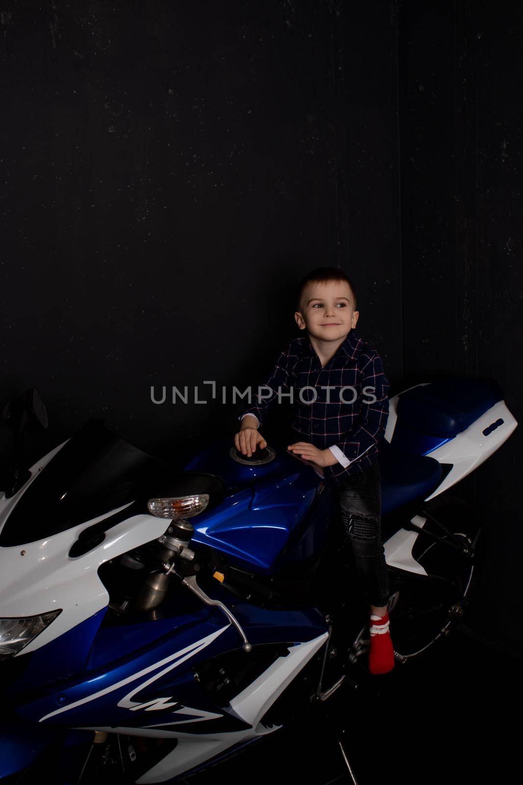 pretty charming little boy on motorcycle on black background. little blonde child riding blue motor bike by oliavesna