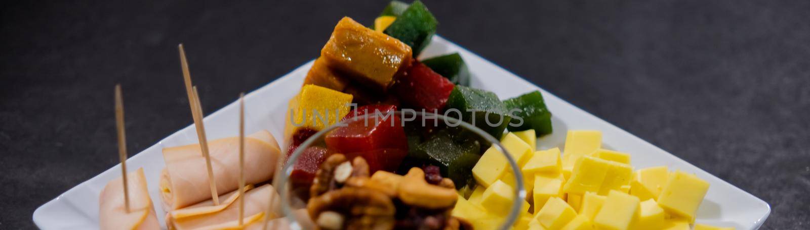 Wide view of turkey ham rolls, diced fruit paste, cheddar cheese cubes, and glass of walnuts on square white plate. Turkey meat, colorful Mexican candy, and nuts above black surface. Healthy snacks