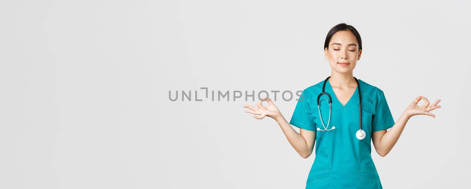 Covid-19, healthcare workers and preventing virus concept. Calm and patient asian female doctor, nurse in scrubs staying relaxed, meditating with eyes closed and happy smile, white background.