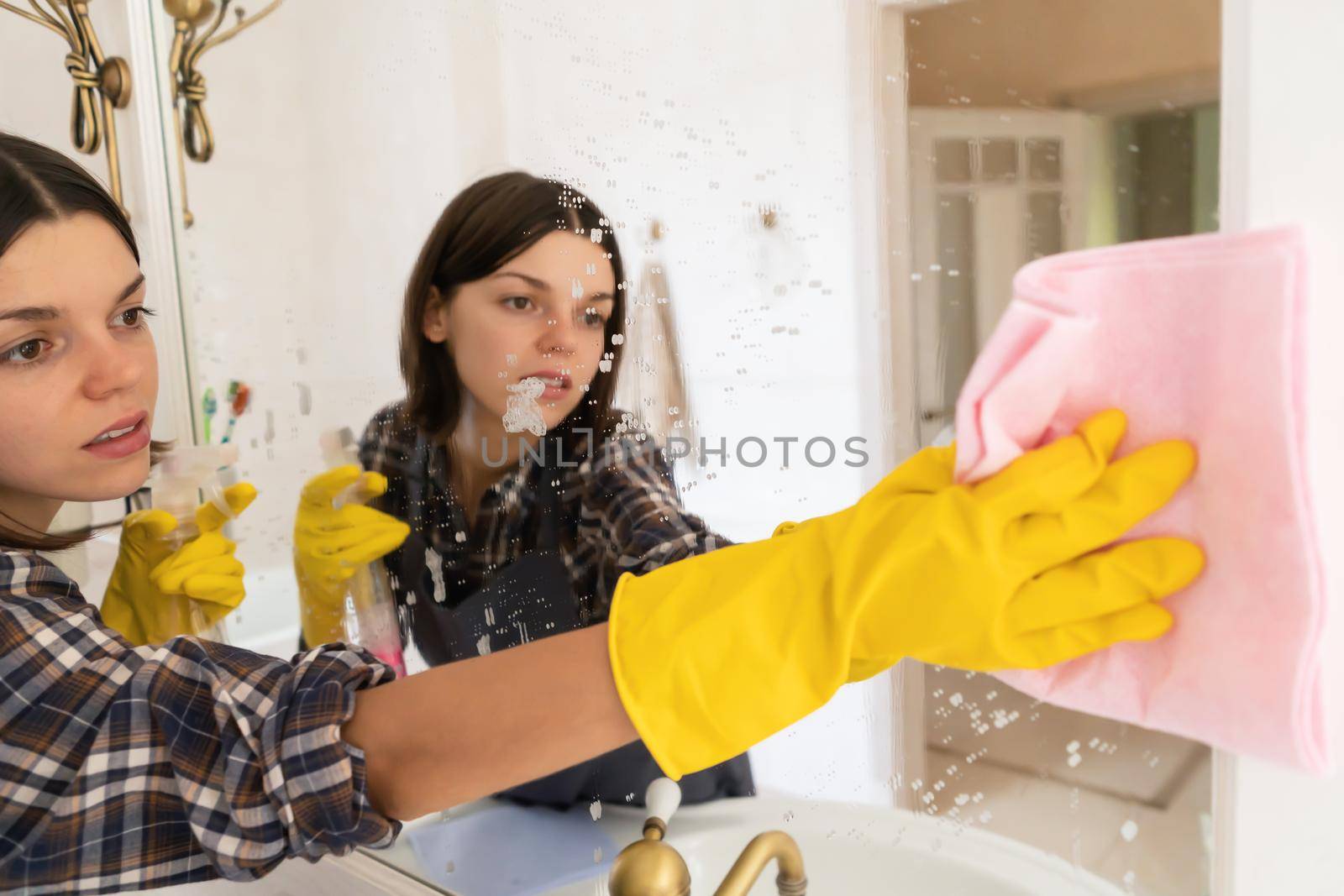 A young girl is cleaning the bathroom, applying detergent with a spray and washing the mirror with a sponge in yellow gloves on her hands. Smiling woman taking care of the cleanliness of her home.