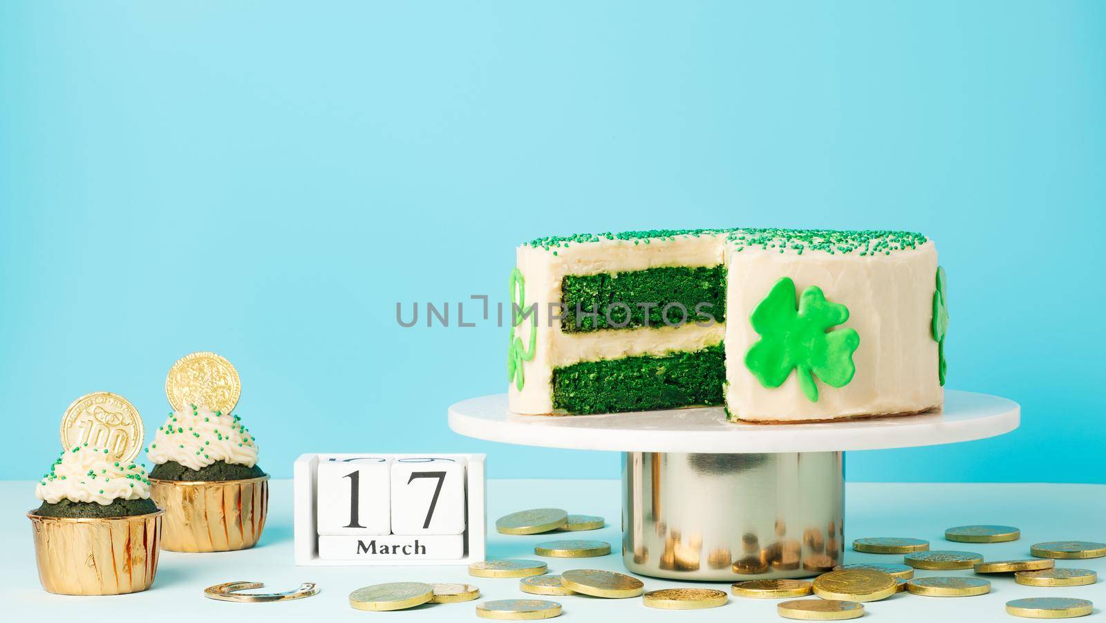 St Patricks Day sweet food concept. Green velvet cake decorated green shamrock leaves and cupcakes with golden coins for Saint Patrick's Day party on blue background. Copy space.Long horizontal banner