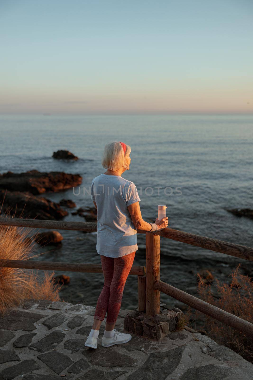 Back view of meditative female pensioner in sportswear, looking at seascape and resting after jogging outdoors