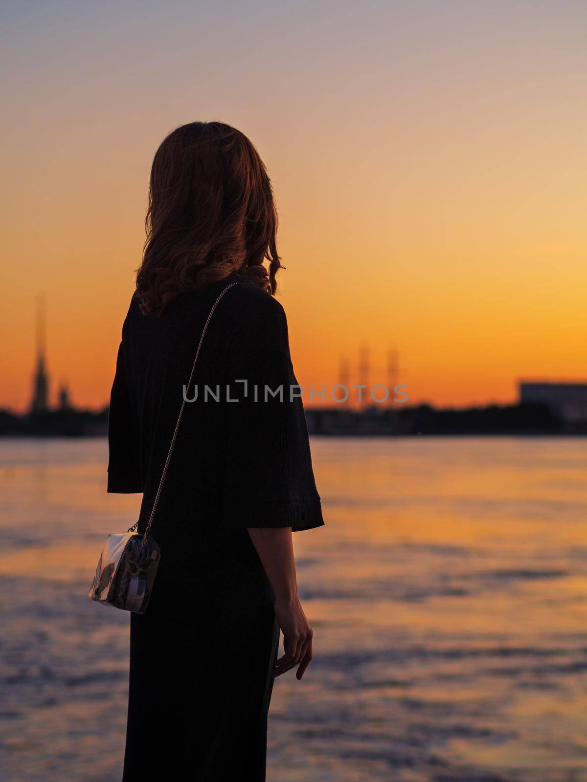 A young elegant woman stands on the embankment and looks at the orange sunset on the Neva River and the Peter and Paul Fortress. St. petersburg by Andre1ns
