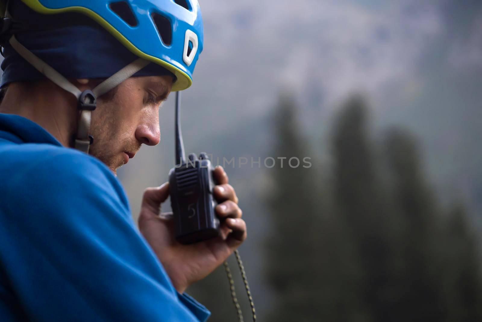 A man in a helmet and a windbreaker is talking on a walkie-talkie against the background of the mountains, a mountain rescuer checks the connection with the base, close-up view.