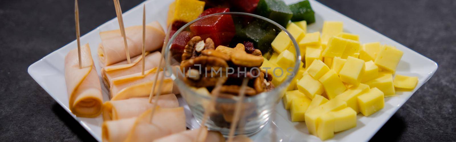 Wide view of turkey ham rolls, diced fruit paste, cheddar cheese cubes, and glass of walnuts on square white plate. Turkey meat, colorful Mexican candy, and nuts above black surface. Healthy snacks