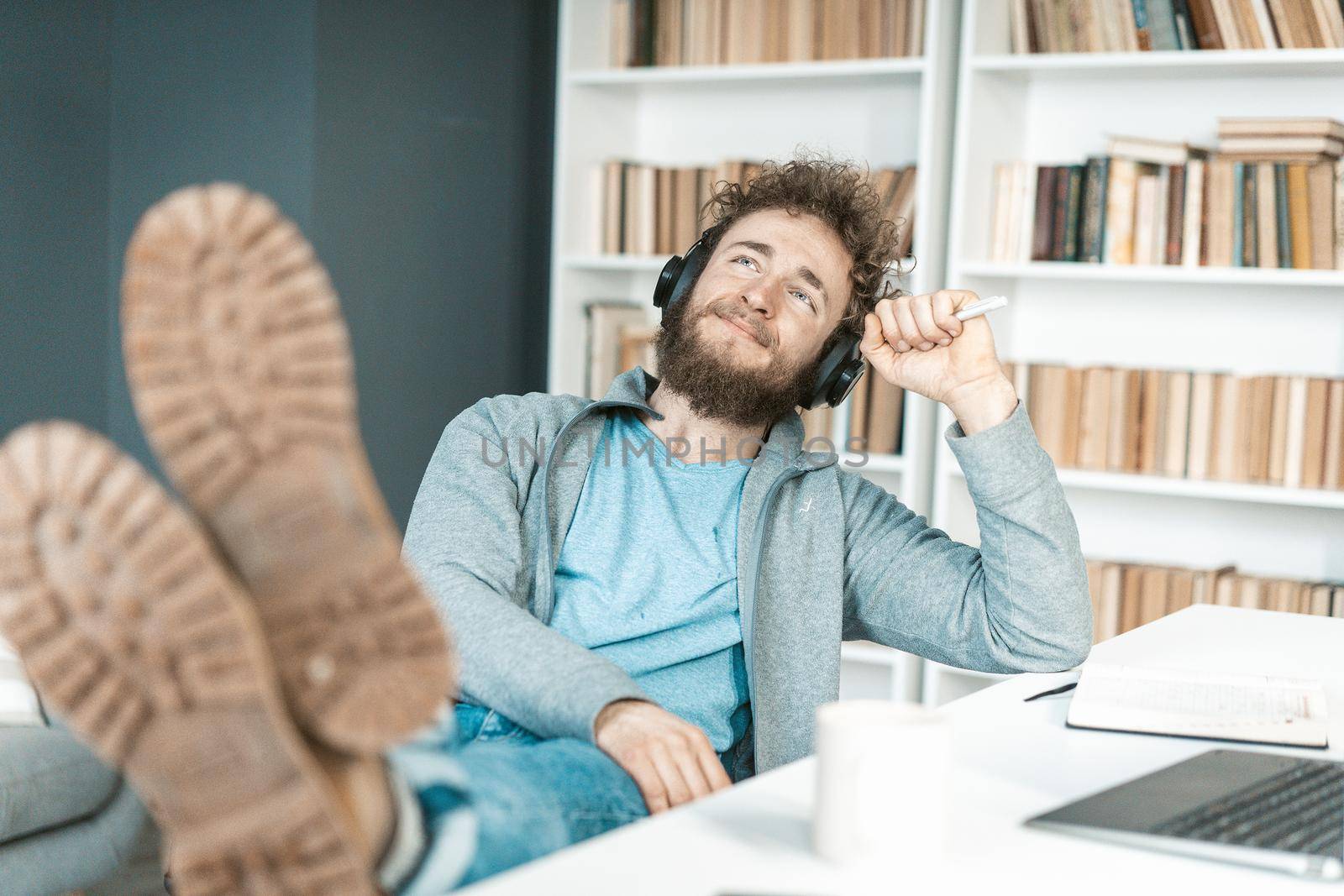Casual Guy Listens to Relaxing Music on Headphones in his Office, Feet on the Table, Soles to the Camera. Bookshelves Background. Close-up. High quality photo