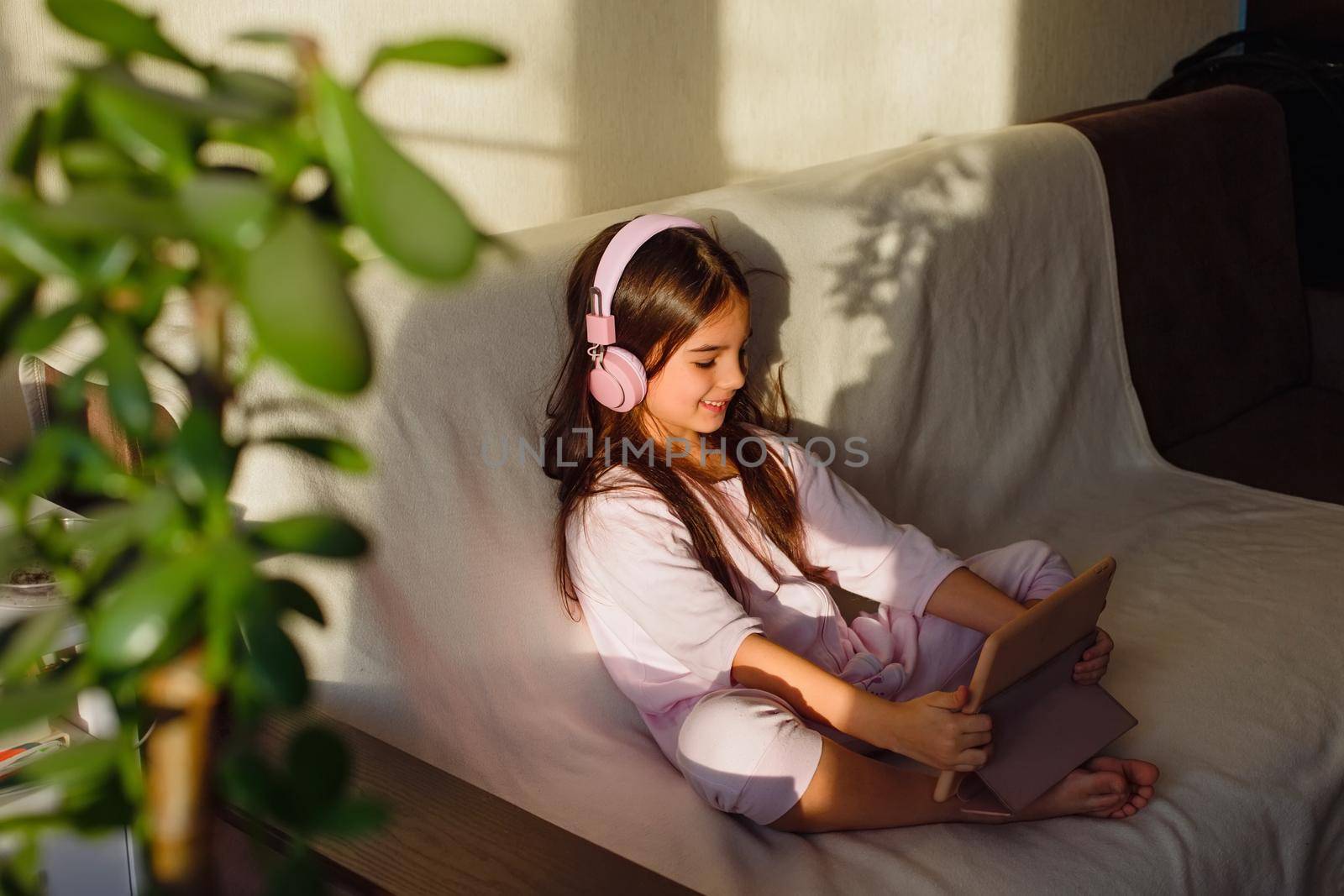 A little smiling girl in cozy pink home clothes and pink headphones sits on the couch, looks into a digital tablet.