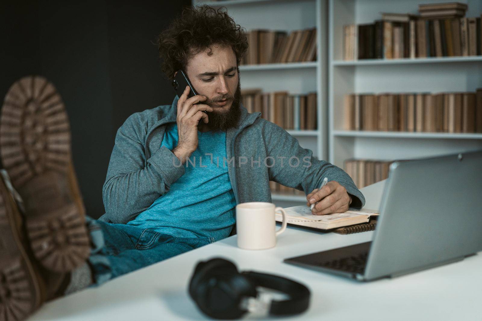 Caucasian Man has a Telephone Conversation and Takes Notes in his Diary. Feet on the Table. Soles to the Camera. Shelves Background. Close-up. High quality photo