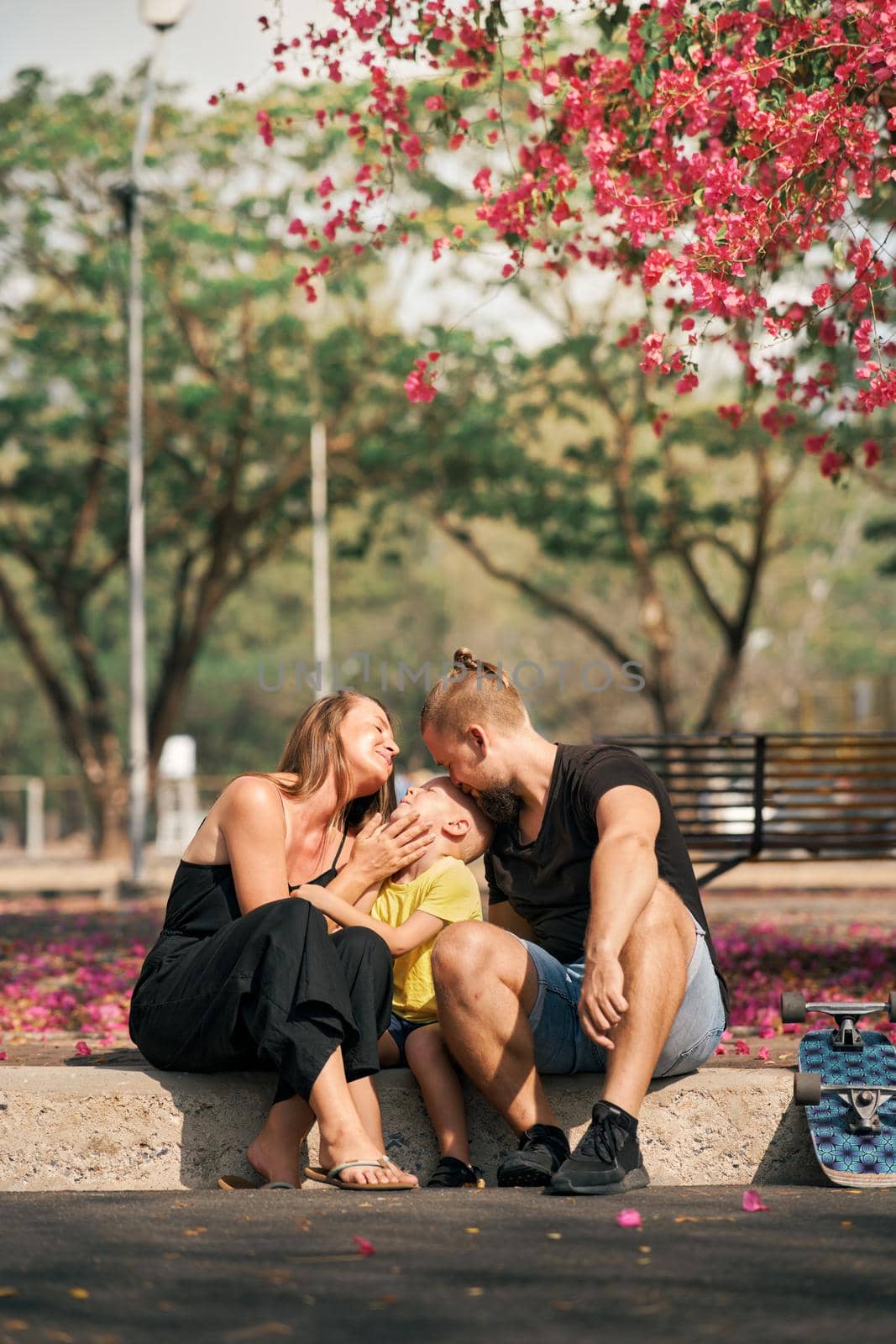 Father and mother is hugging their little son while sitting in skate park with blossom trees around. Concept of family love and care. Outdoor leisure and activity. Happy family.