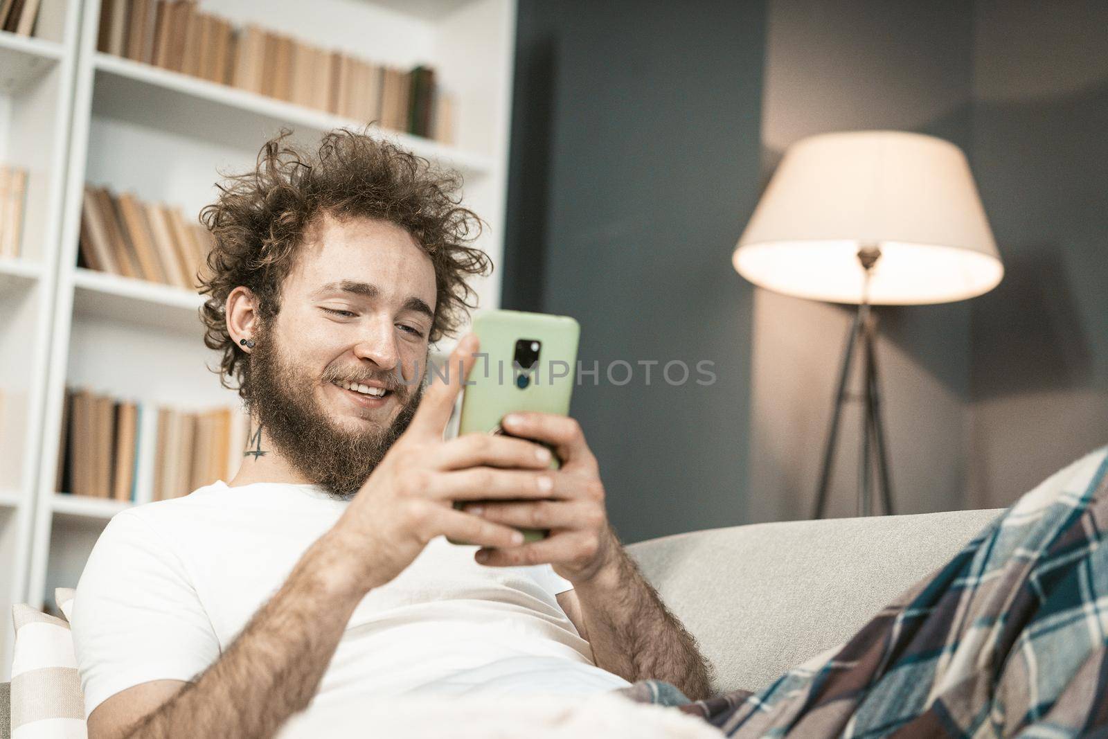 Tired Guy Lies in his Bed and Plays Relax Game on his Smartphone Before Bedtime. Exhausted Student Reads the News on his Mobile at Night. Home Decor Background. Close-up. High quality photo