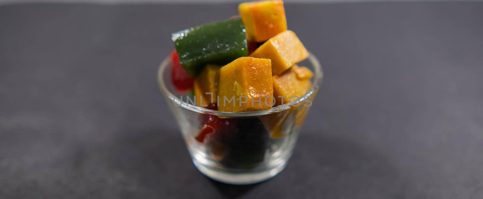 Wide view of glass of red, yellow, and green fruit paste slices on black surface. Close-up of authentic natural Mexican candy cubes in glass cup. Tasty traditional snacks